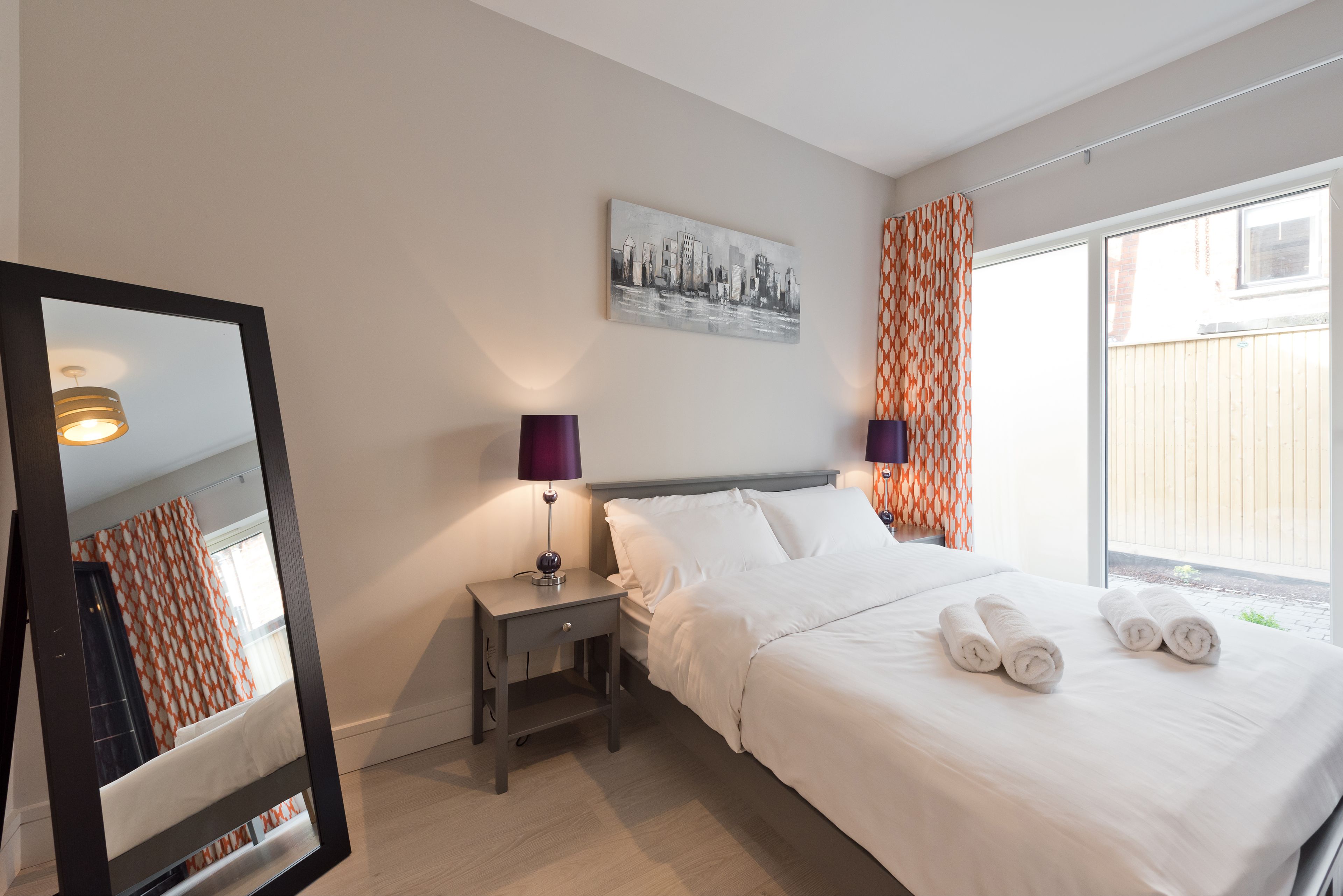 Une chambre d’hôtel avec un lit, une table de chevet avec une lampe, un miroir et un tableau représentant un paysage urbain accroché au mur.