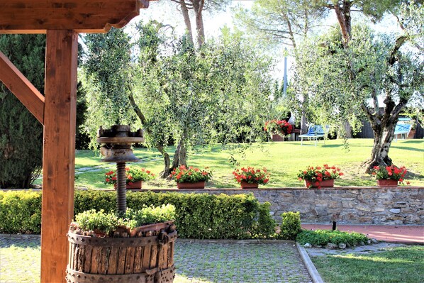 A wooden pergola with a rustic bird feeder, surrounded by a stone wall and potted red flowers.