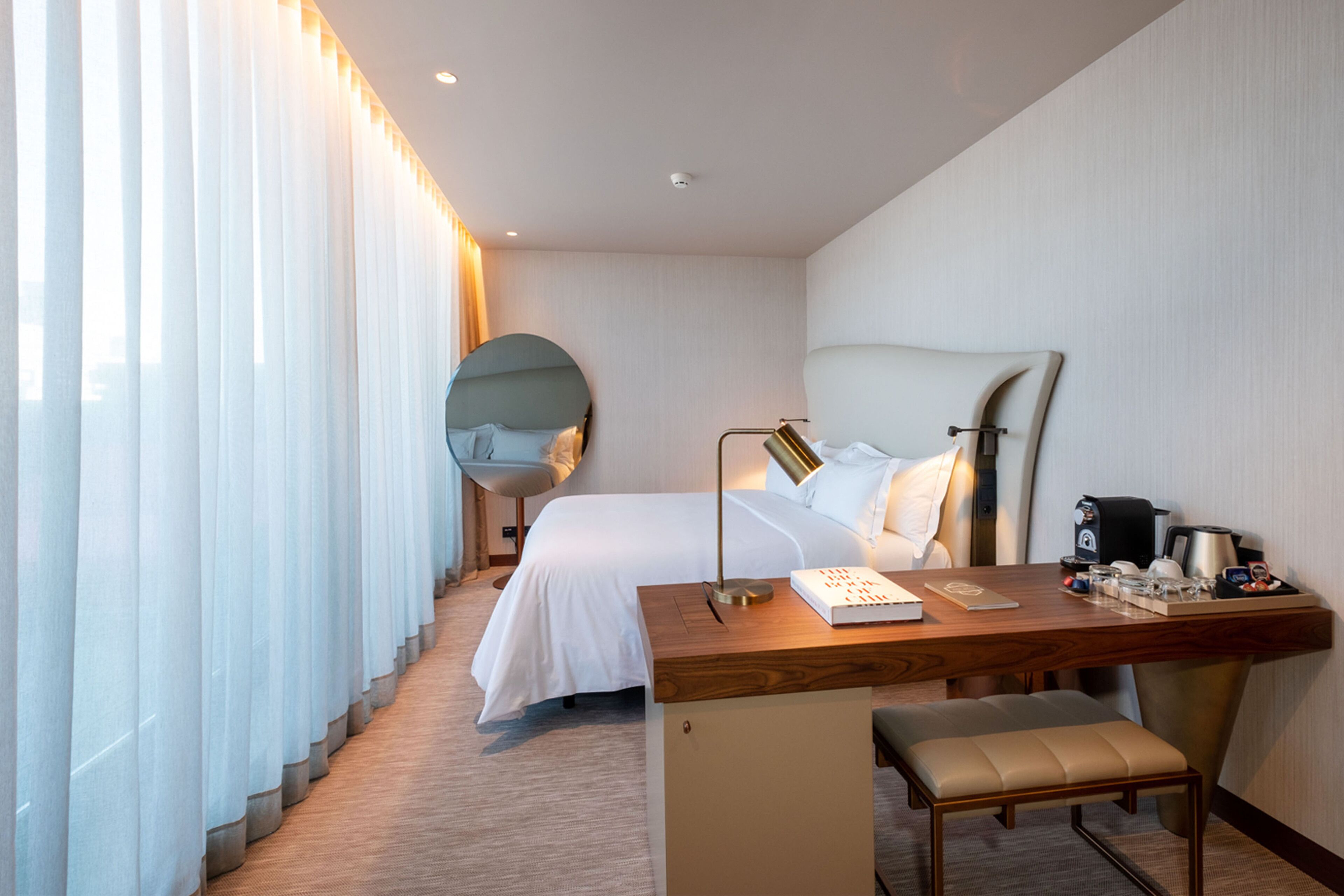 A hotel room with a bed, a desk with a lamp, a coffee maker, and a chair.
