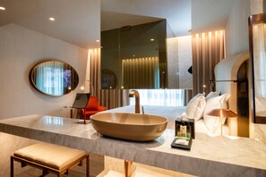 Un baño moderno con un lavamanos ovalado grande, un espejo redondo, un banco y un mueble de tocador con artículos de aseo.