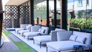 A modern outdoor seating area with sofas and tables, a wooden deck, and a view of greenery and buildings.