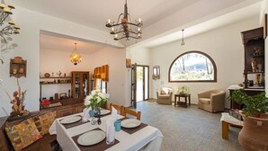 A spacious living area with a dining table set for four, a chandelier, and a large arched window providing a view of the outdoors.
