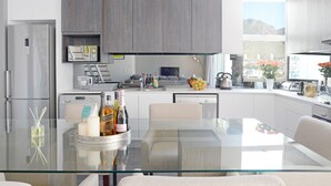 A modern kitchen with a glass dining table, grey cabinets, and a view of mountains through the window. - Naomi By Stay Amazing (Cape Town)