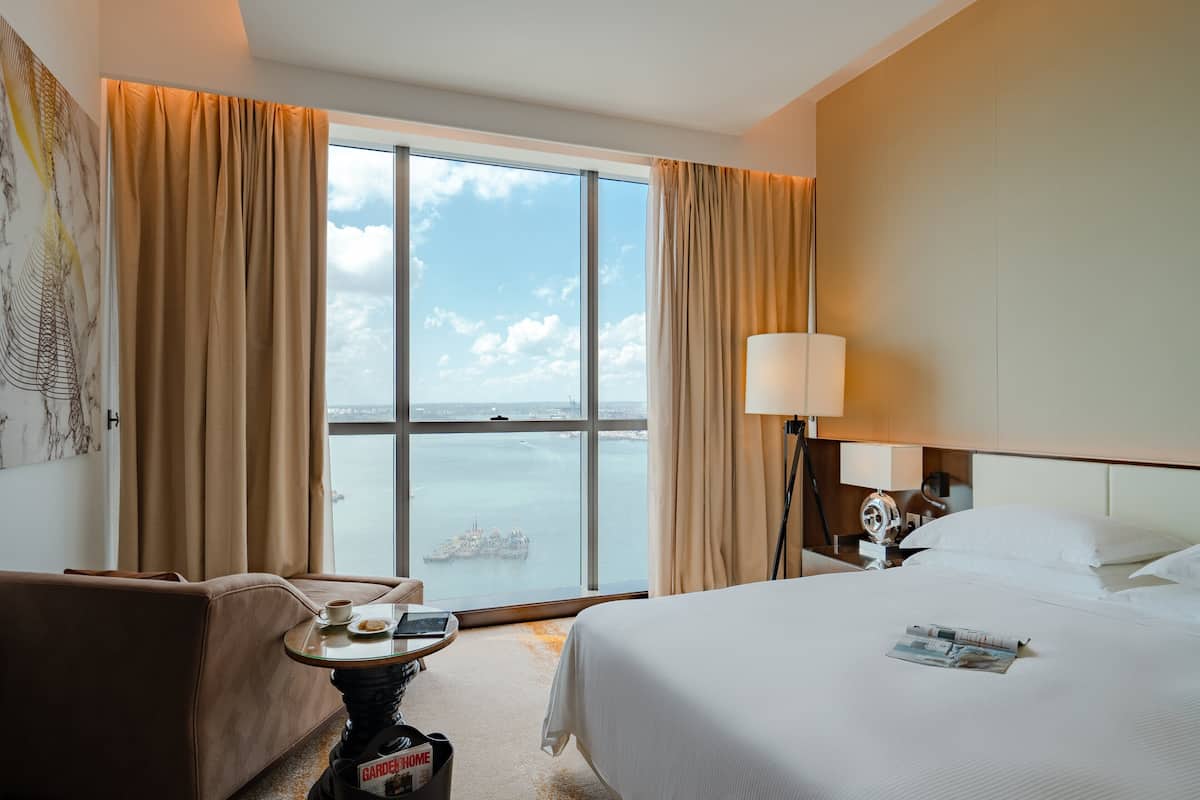 A hotel room with a large window, a bed, a bedside table with a lamp, and a view of the sea.