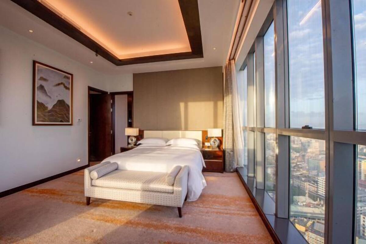 A hotel room with a large bed, a city view through floor-to-ceiling windows, a painting on the wall, and a ceiling with recessed lighting.
