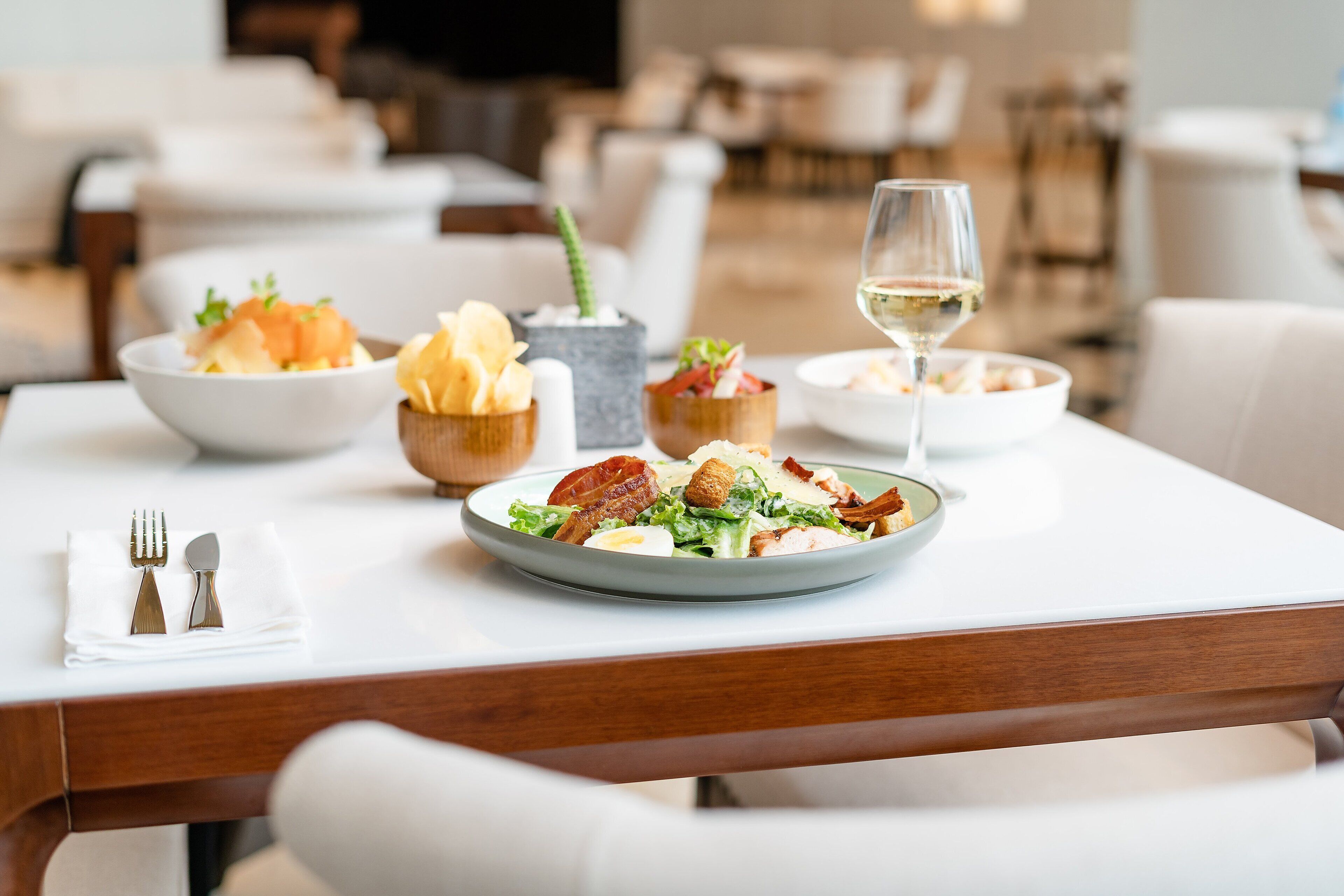 Una mesa de comedor con una ensalada, una copa de vino blanco y diferentes acompañamientos.