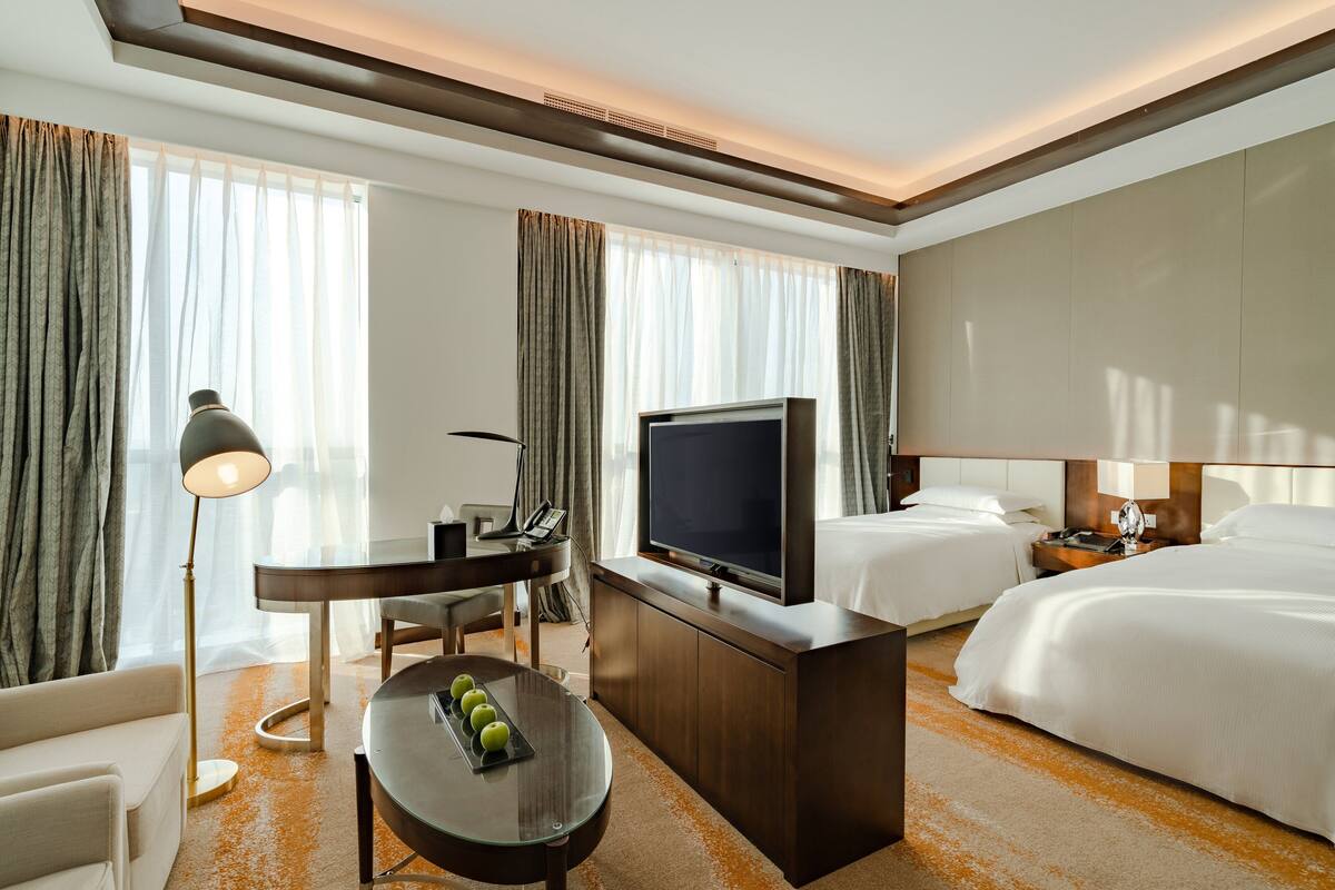 A hotel room with two beds, a TV, a desk with a lamp, and a dining table with apples.