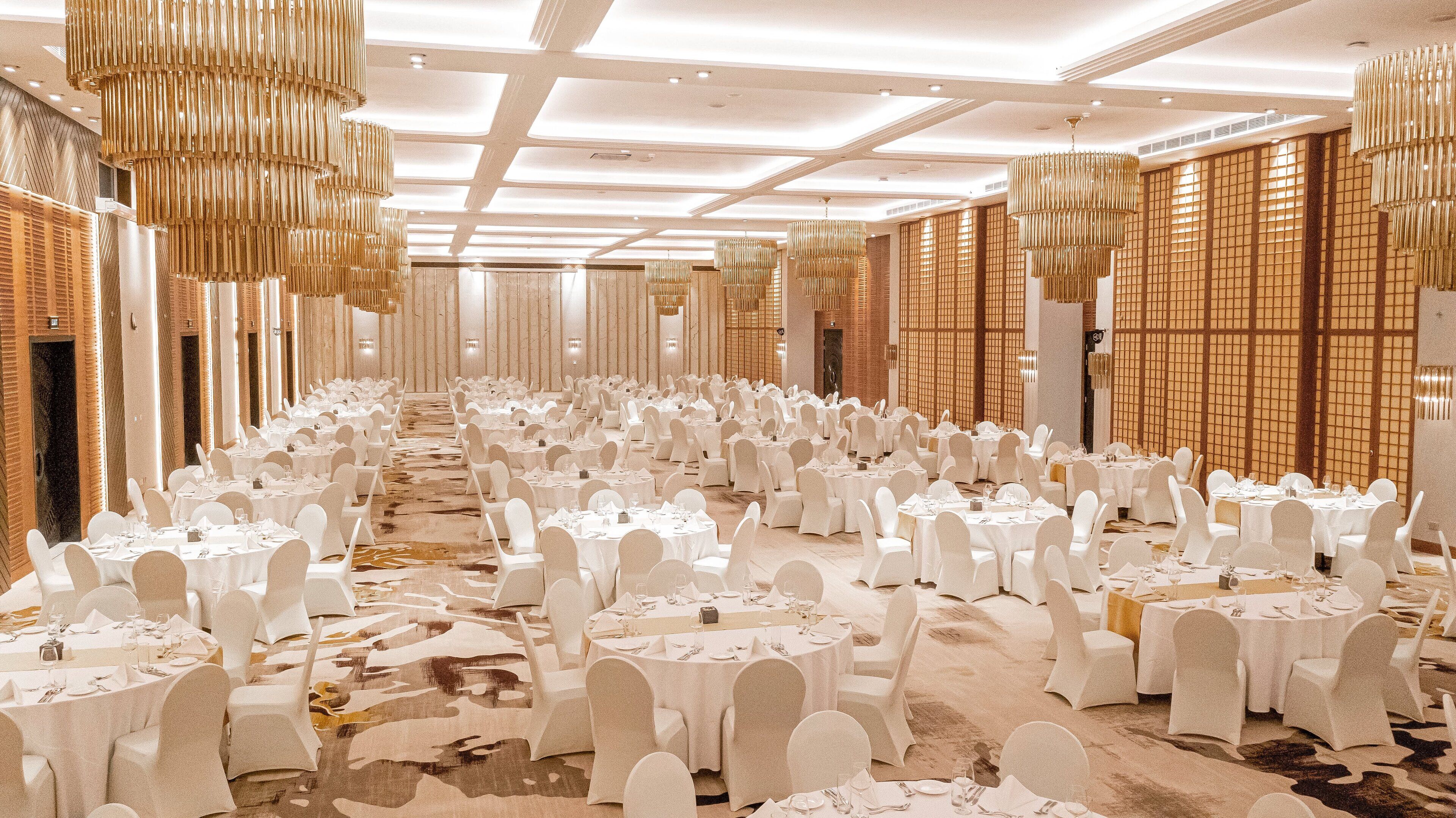 Un salón de banquetes con filas de mesas redondas dispuestas para comer, sillas blancas y lámparas doradas colgantes.