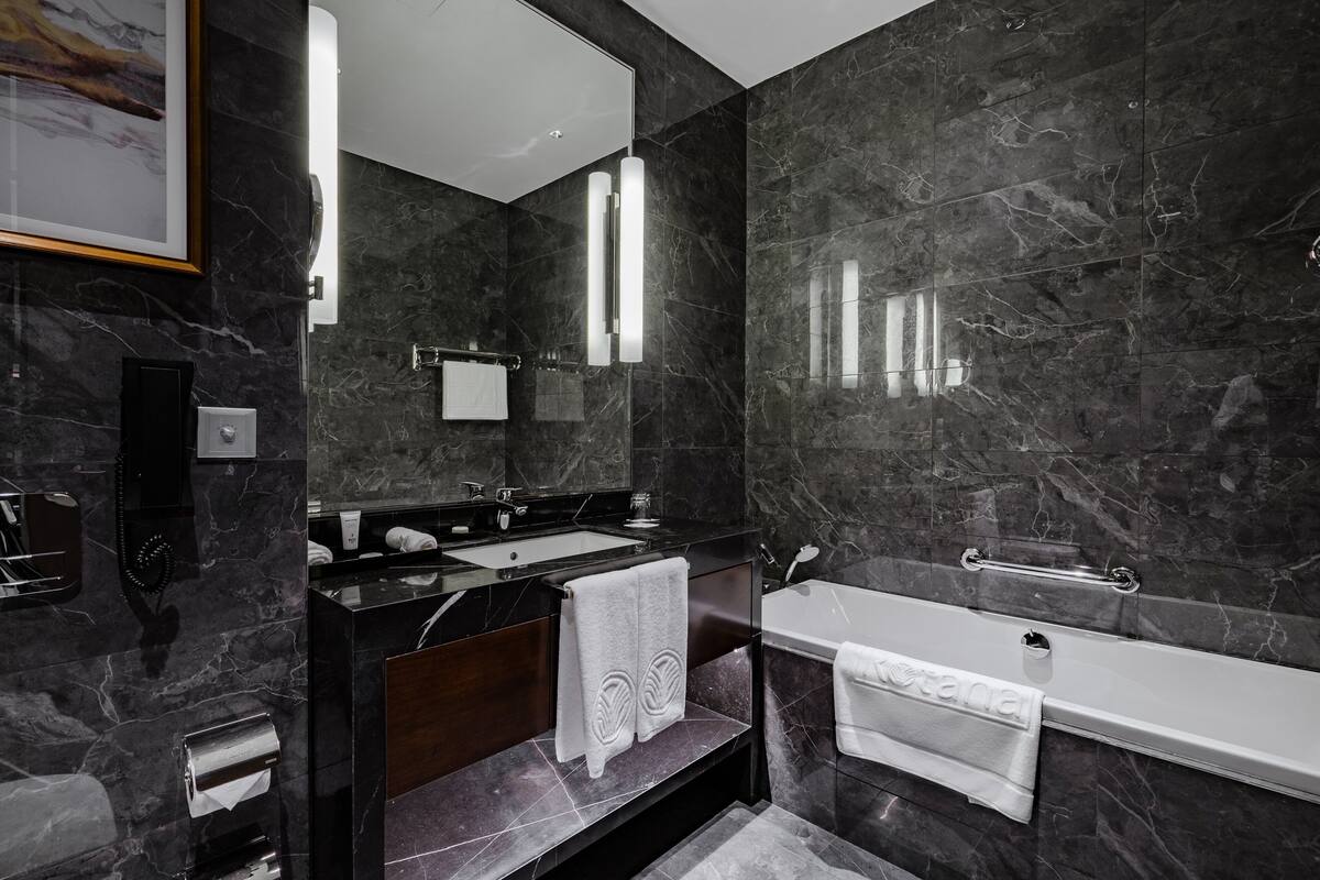 A modern bathroom with dark marble walls, a large mirror, a bathtub, and a vanity with a sink and towel rack.