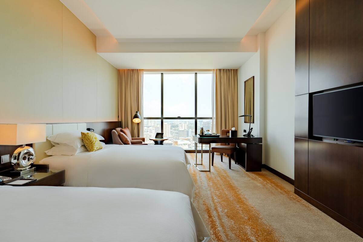 A hotel room with two beds, a desk, a chair, a TV, and a large window with curtains.