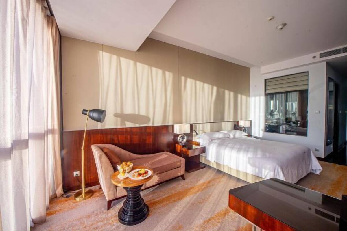 A hotel room with a bed, a sofa, a small table with a plate of food, a lamp, and a large window.