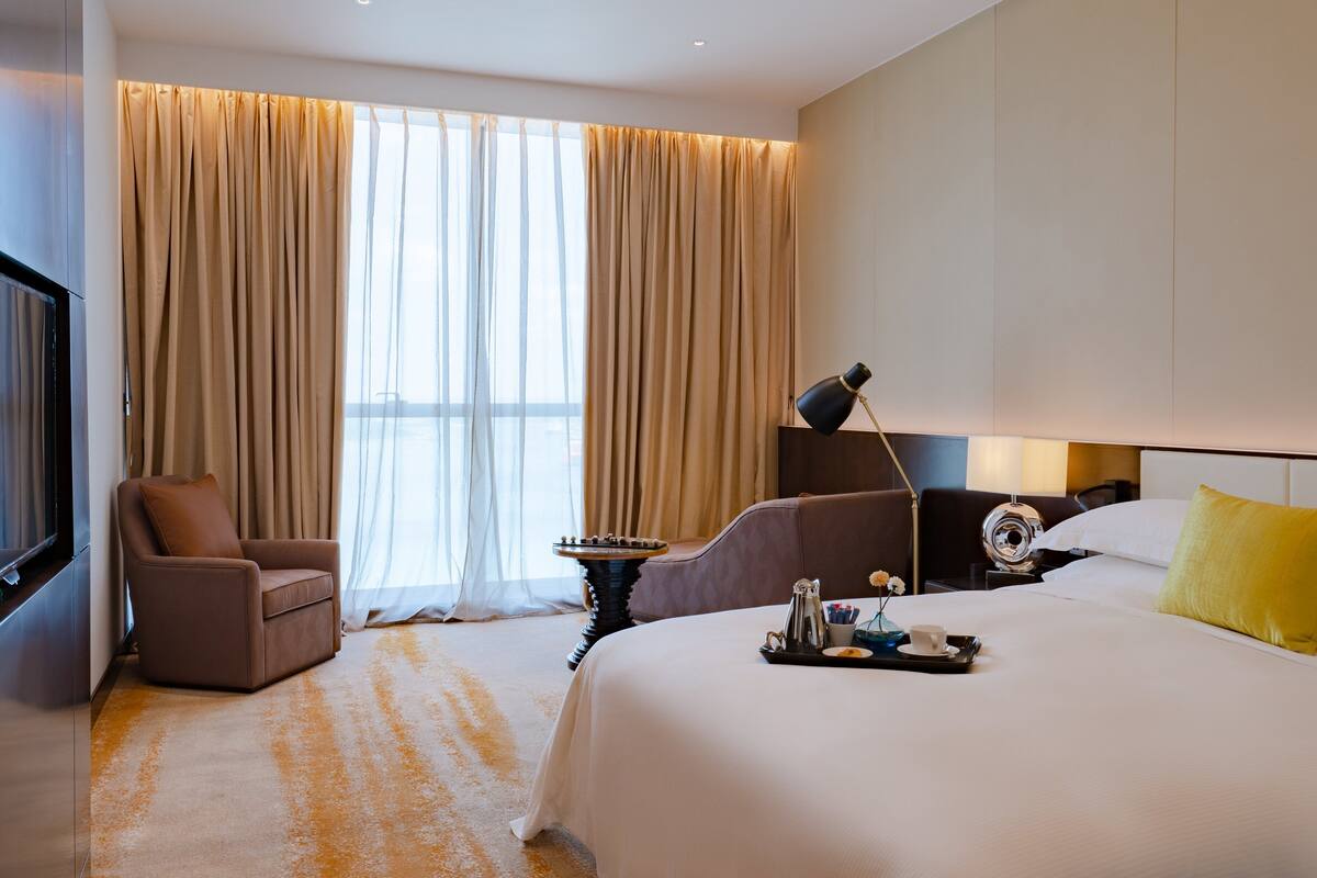 A hotel room with a bed, a chair, a desk with a lamp, and a window with curtains.
