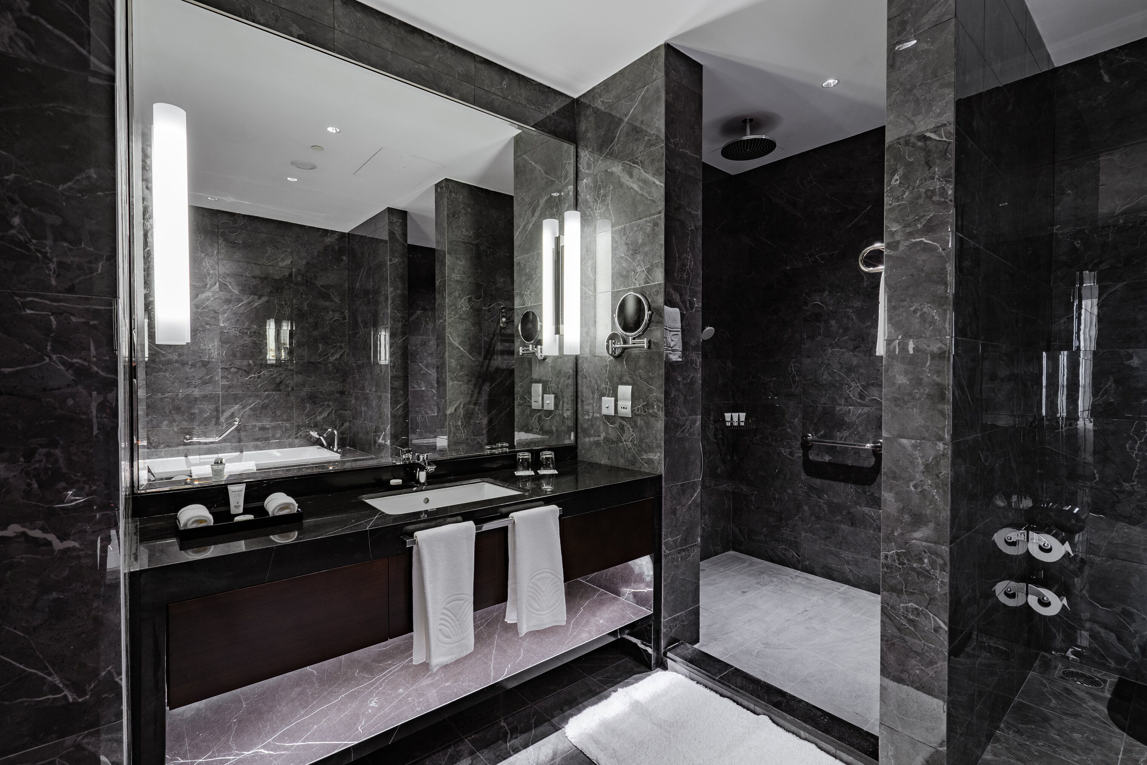 A modern bathroom with a large mirror, dark marble walls, a black countertop, and a glass-enclosed shower area.