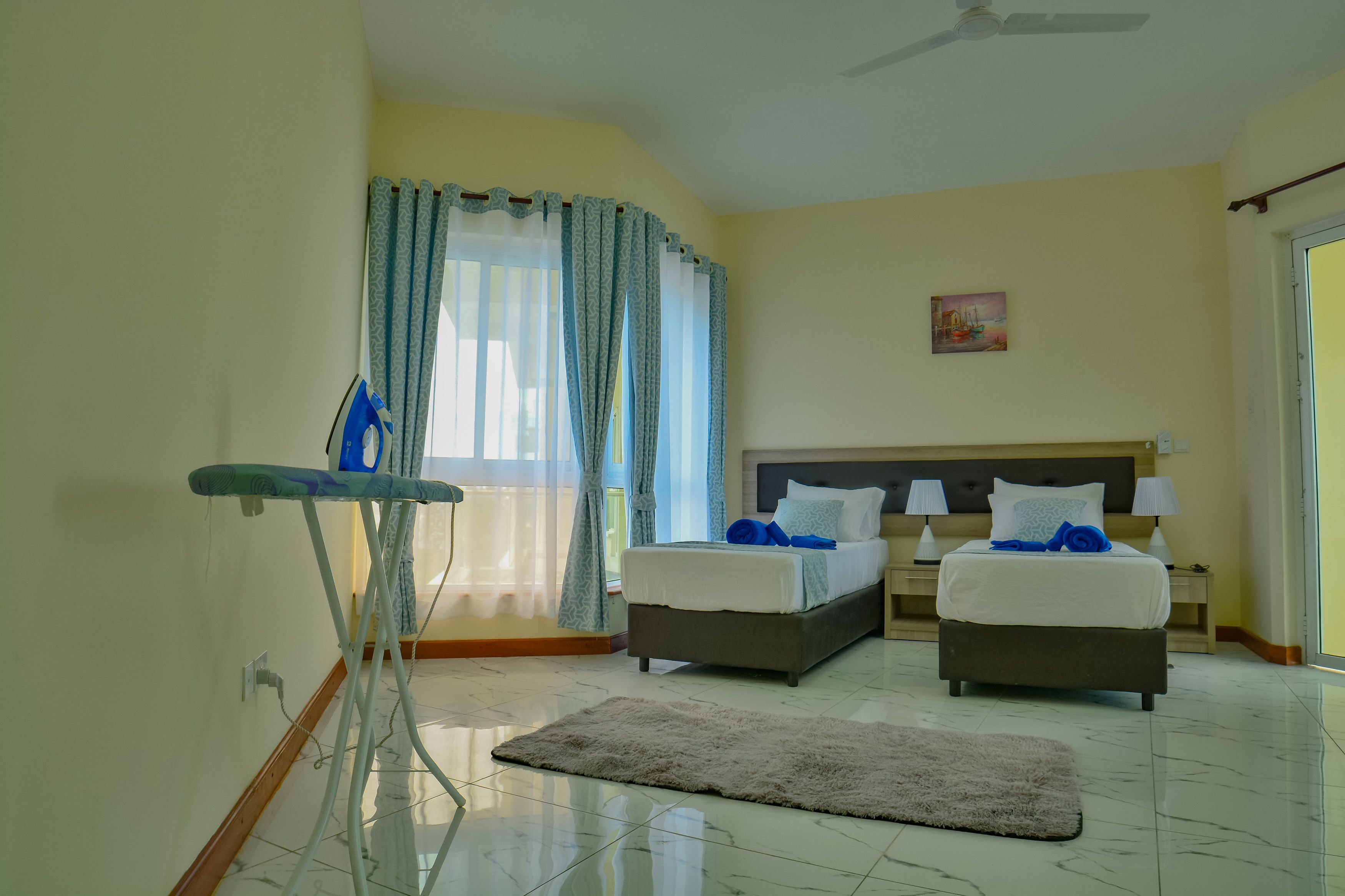 Habitación de hotel con dos camas, ventilador de techo, un cuadro en la pared y una ventana con cortinas.
