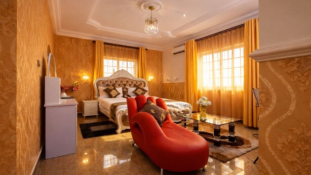 A spacious bedroom with a large bed, a red chaise lounge, a glass coffee table, and a white dresser. - AG Hotel and Suites (Accra)