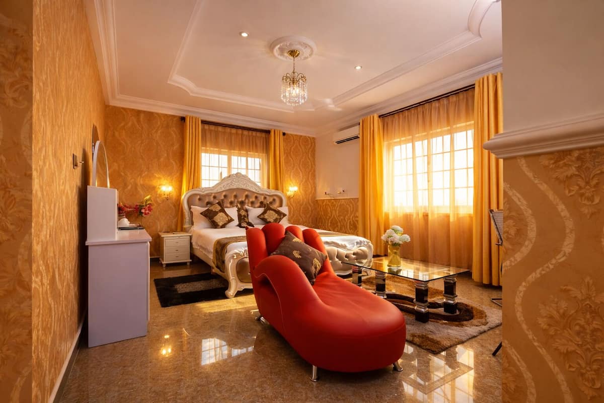 A spacious bedroom with a large bed, a red chaise lounge, a glass coffee table, and a white dresser.
