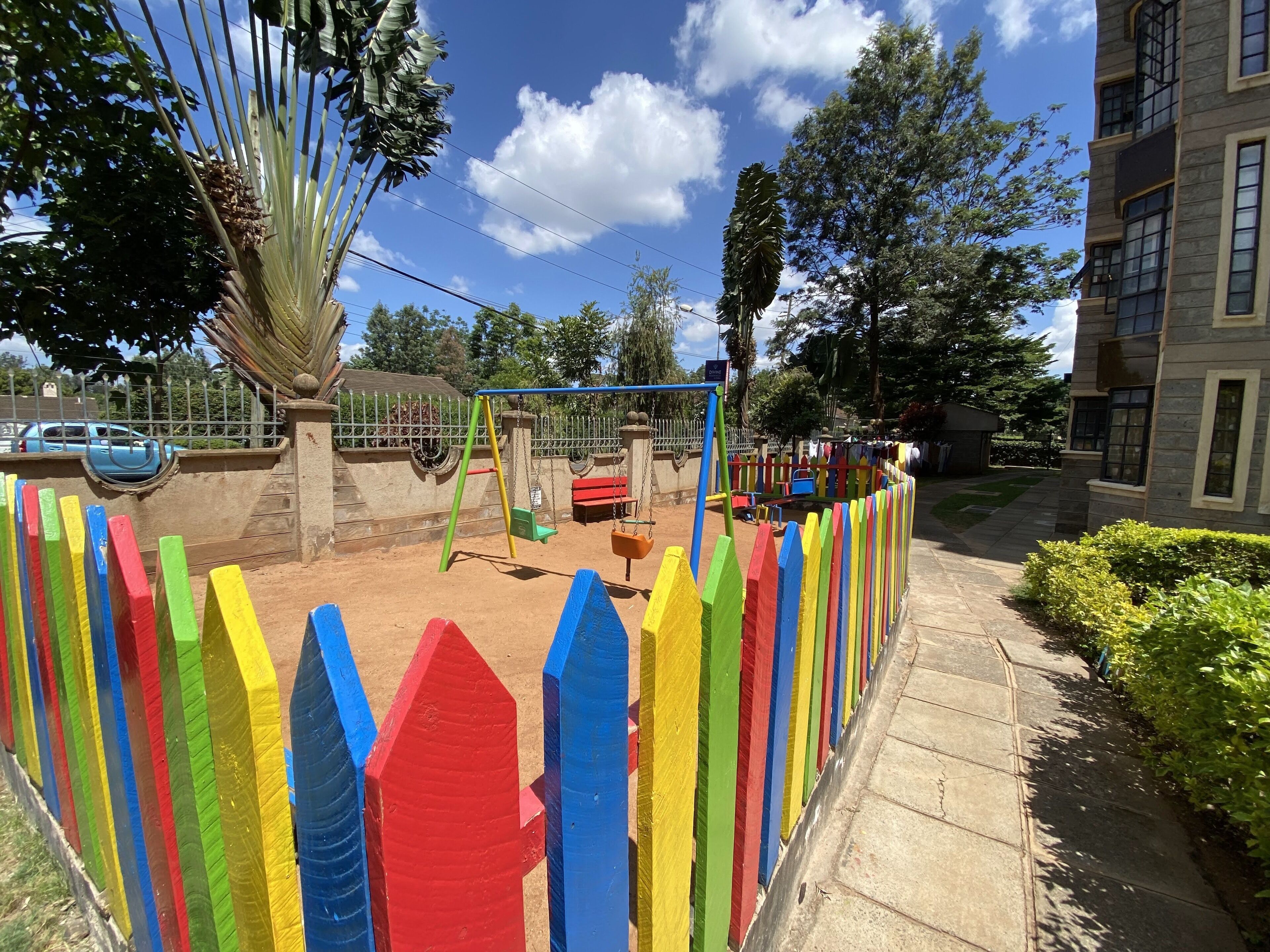 A playground with colorful play structures and a fenced area, surrounded by trees and a building.