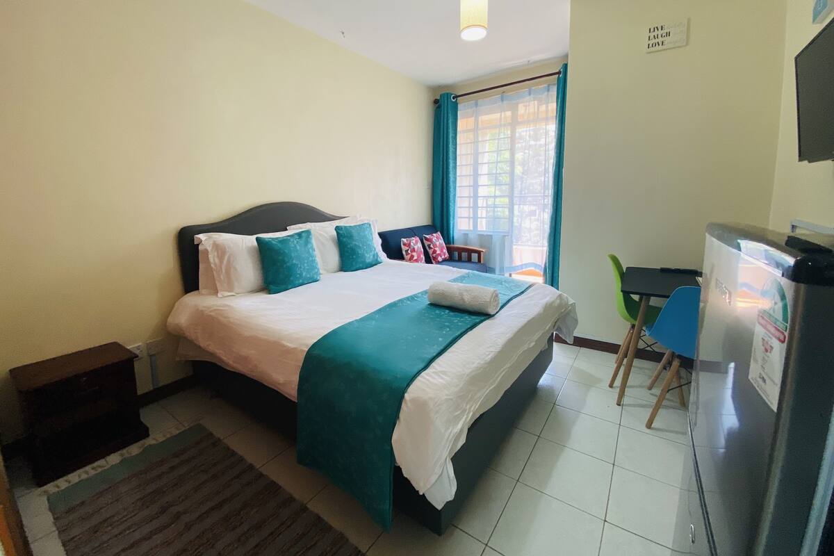Un dormitorio con cama, escritorio, silla, refrigerador y ventana con cortinas.