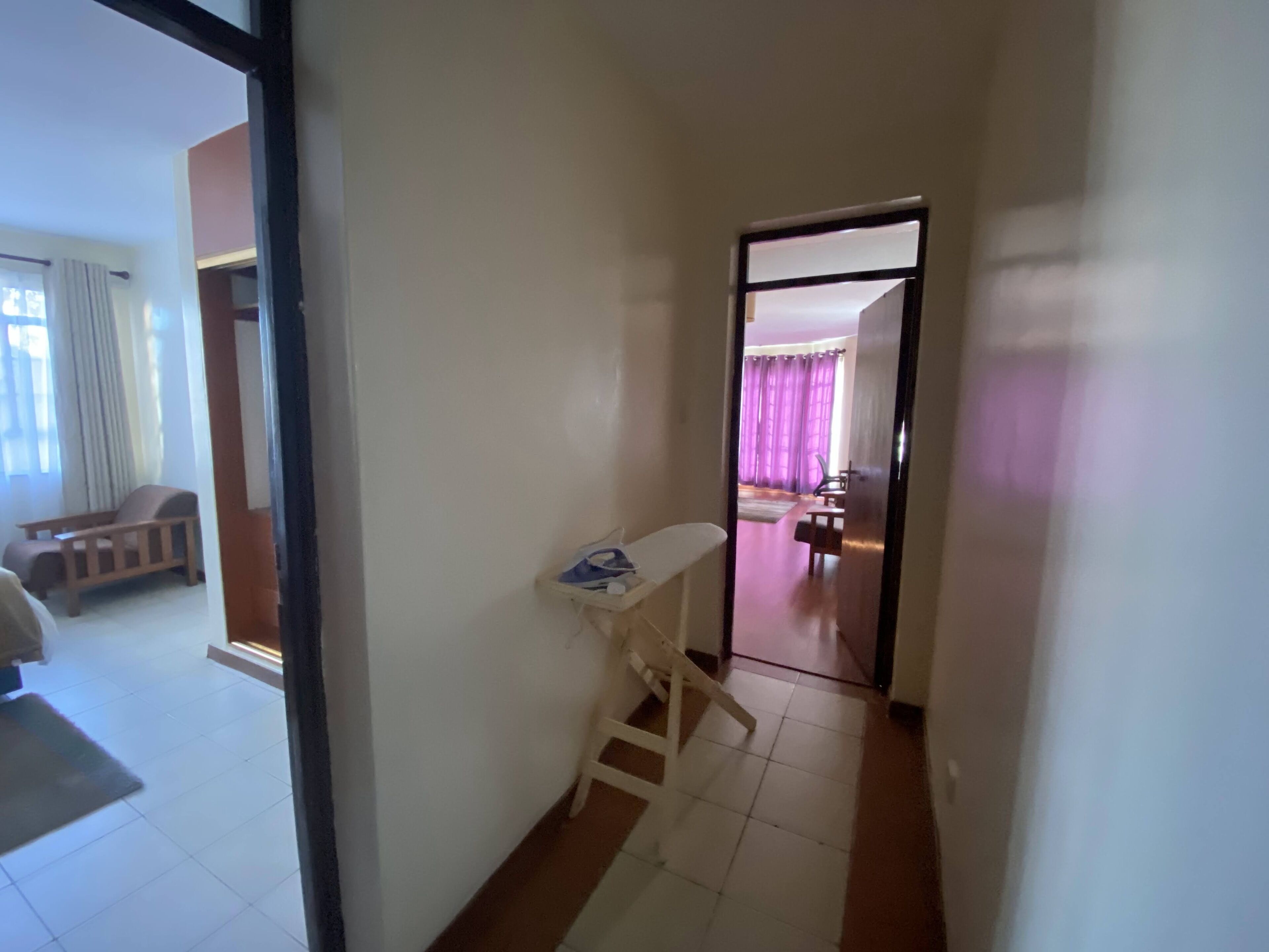 A hallway with a white chair and a small table, leading to a room with a bed and a window with curtains.