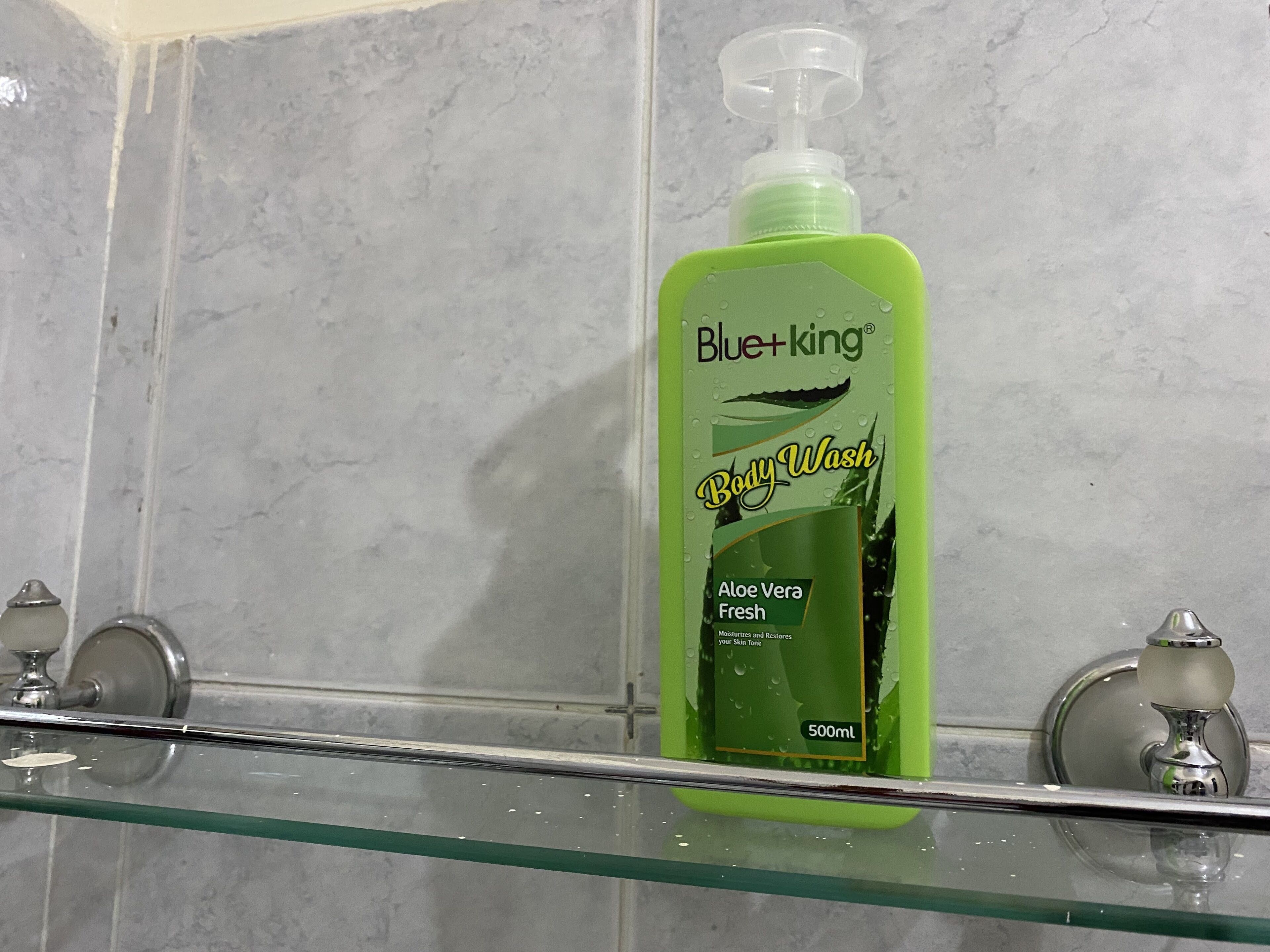 Casa de banho com cabine de chuveiro em vidro, um frasco verde de gel de banho Blue+king Aloe Vera Fresh e uma parede de mármore.