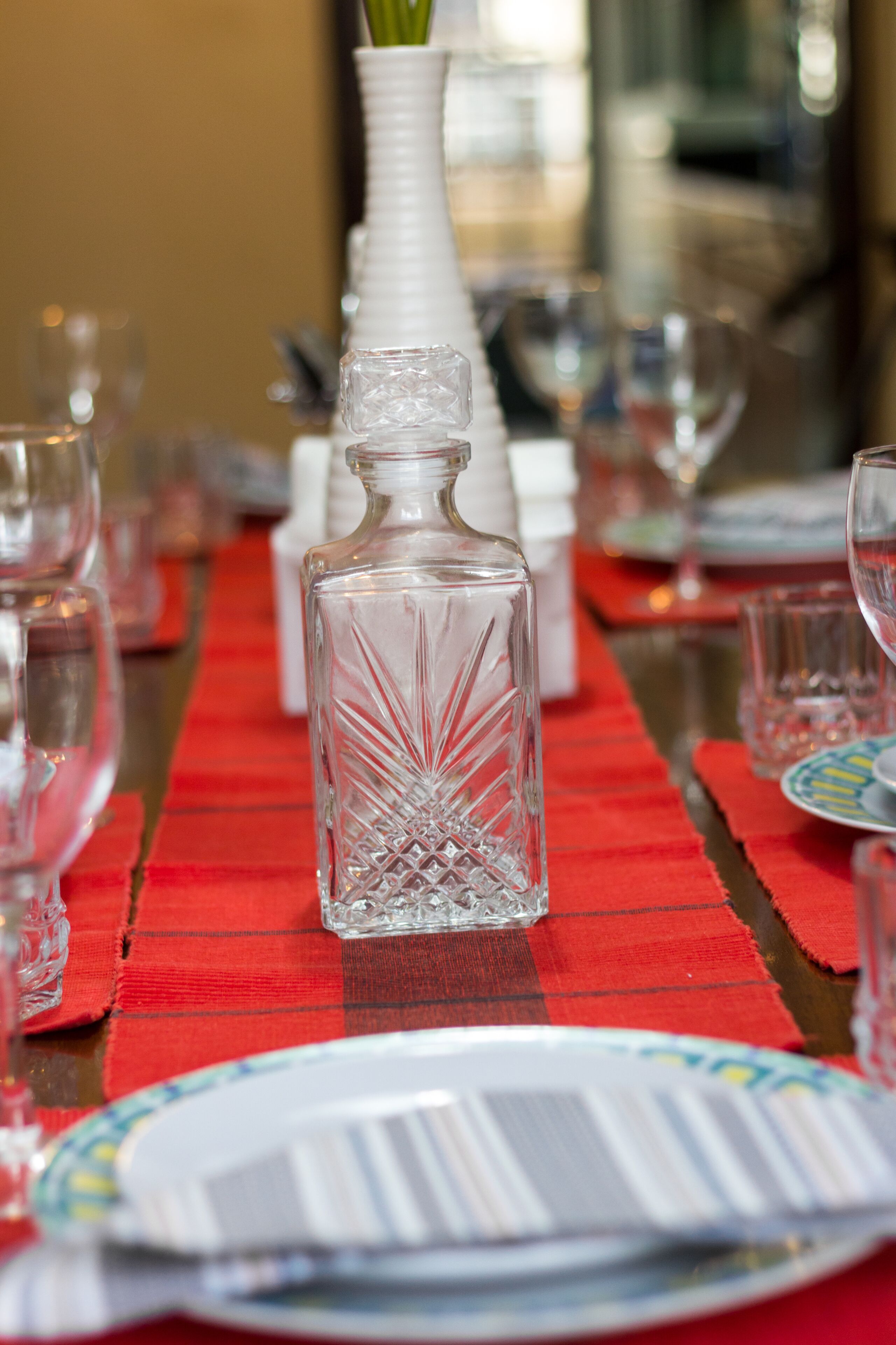 Una mesa de comedor dispuesta con cristalería transparente, un florero blanco con una planta verde y un mantel individual rojo.