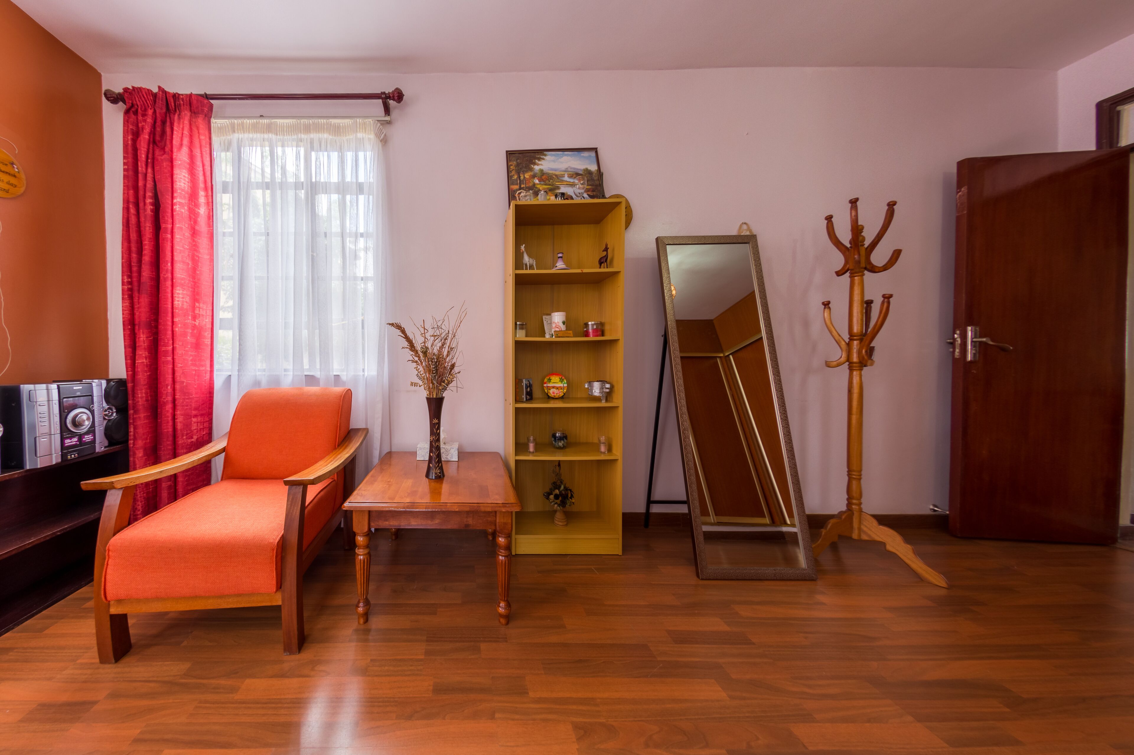 Une pièce avec un plancher en bois, une chaise rouge, une table en bois, une bibliothèque jaune, un miroir et un porte-manteau en bois.