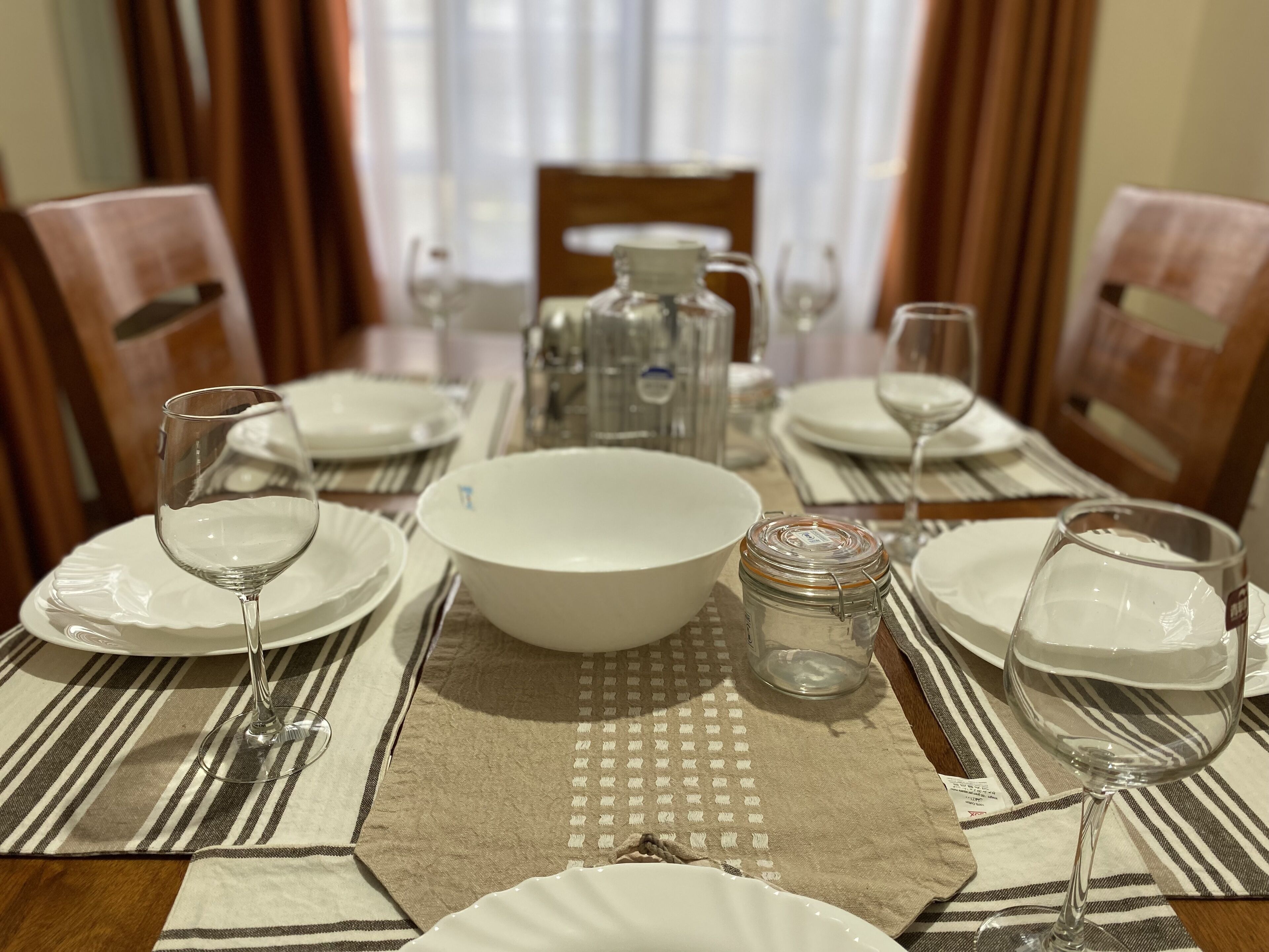 A dining table set with white plates, bowls, and wine glasses, with a striped placemat and a clear glass jar in the center.
