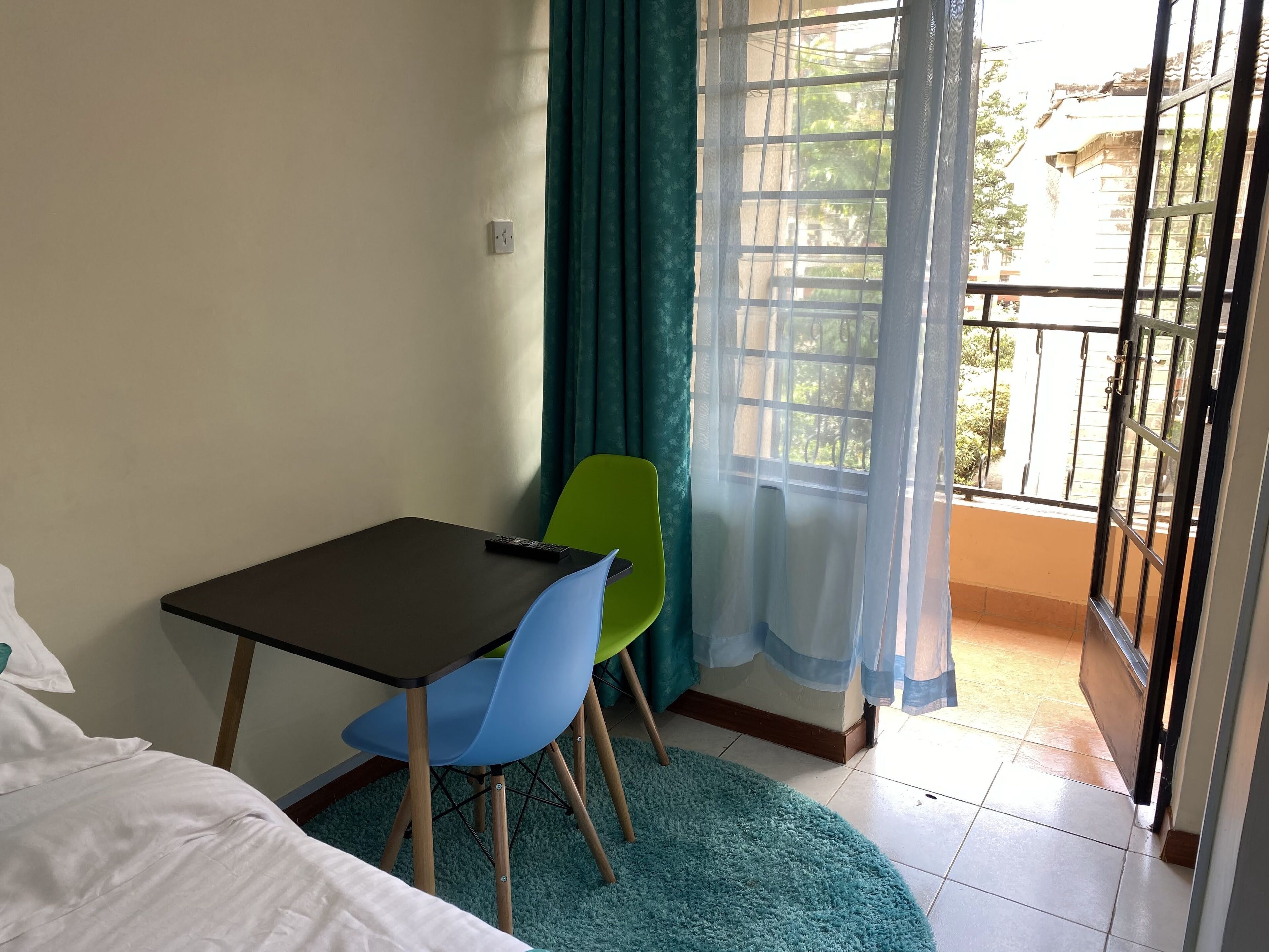 A small room with a bed, a table, and a chair. There is a window with curtains and a view of the outside.