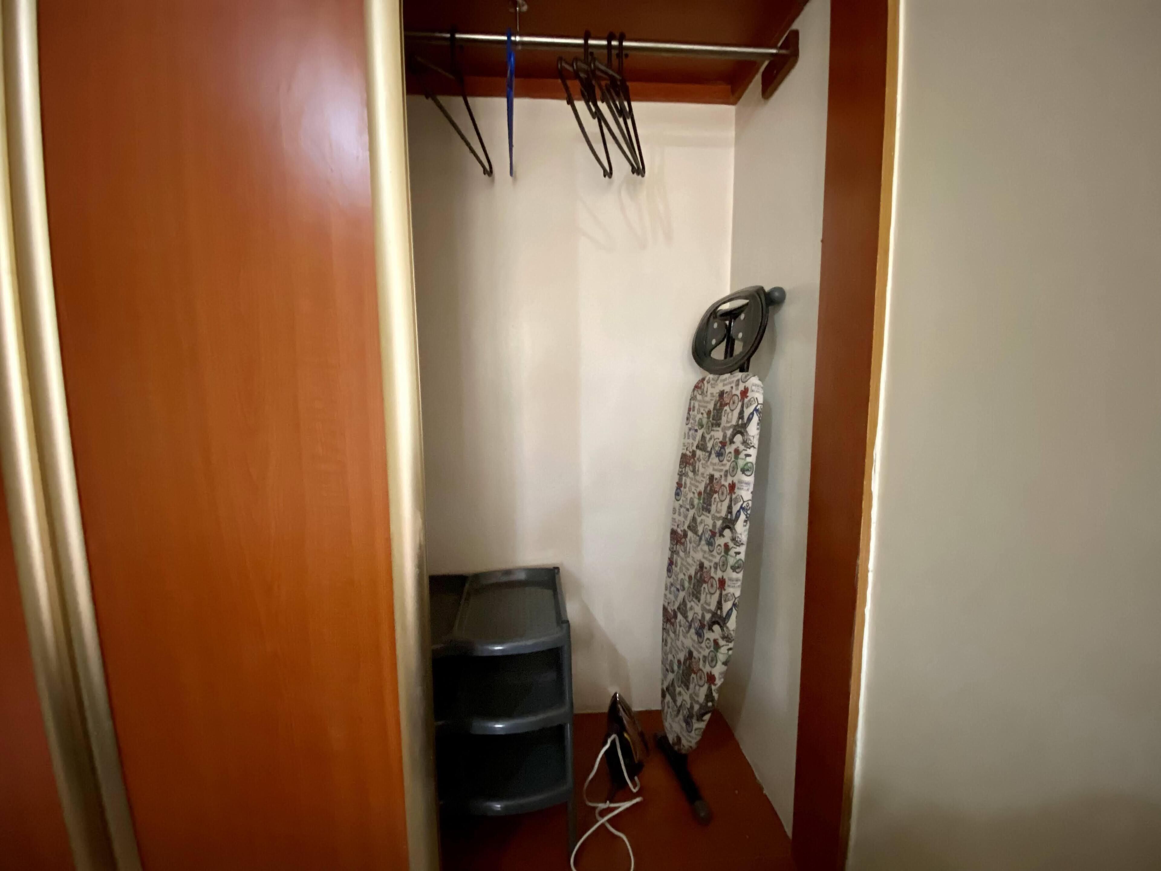 A small, narrow room with a wooden door, a drying rack, a patterned curtain, and a black chair.