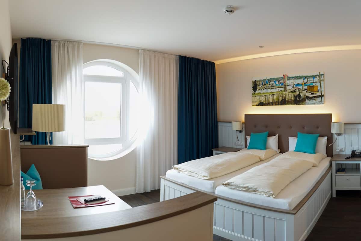 Ein Hotelzimmer mit zwei Betten, einem Schreibtisch, einem Sessel, einer Lampe, einem Gemälde und einem Fenster mit Vorhängen.