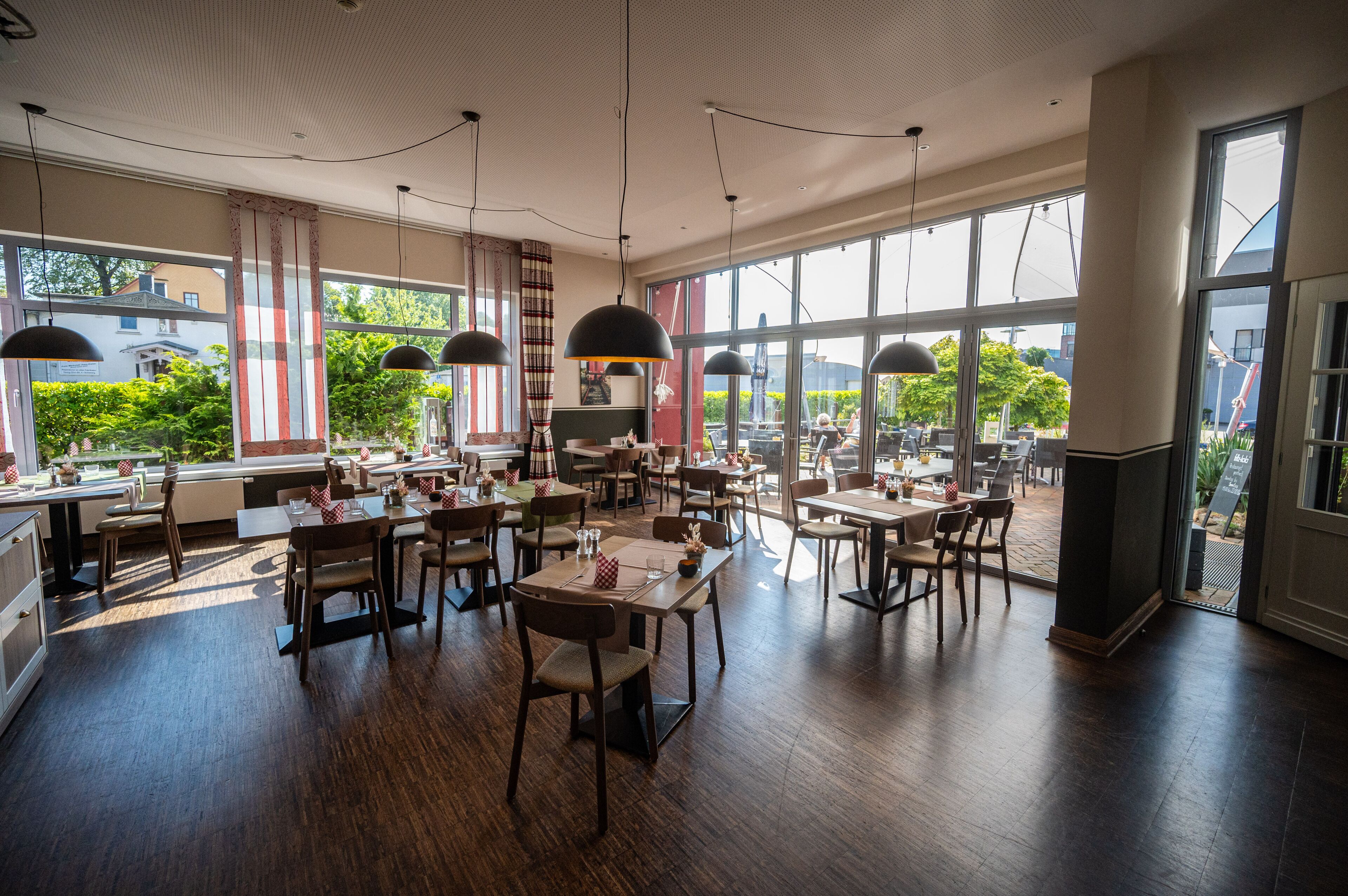 A spacious restaurant with high ceilings, large windows, and modern pendant lighting. Tables are neatly arranged with chairs, and the interior features a mix of wood and metal elements.