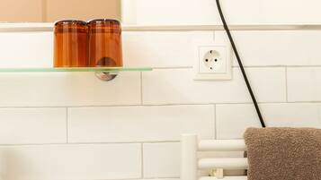 A bathroom with a white bathtub, a towel rack with a brown towel, a hairdryer on a wall socket, and two amber-colored bottles on a glass shelf.