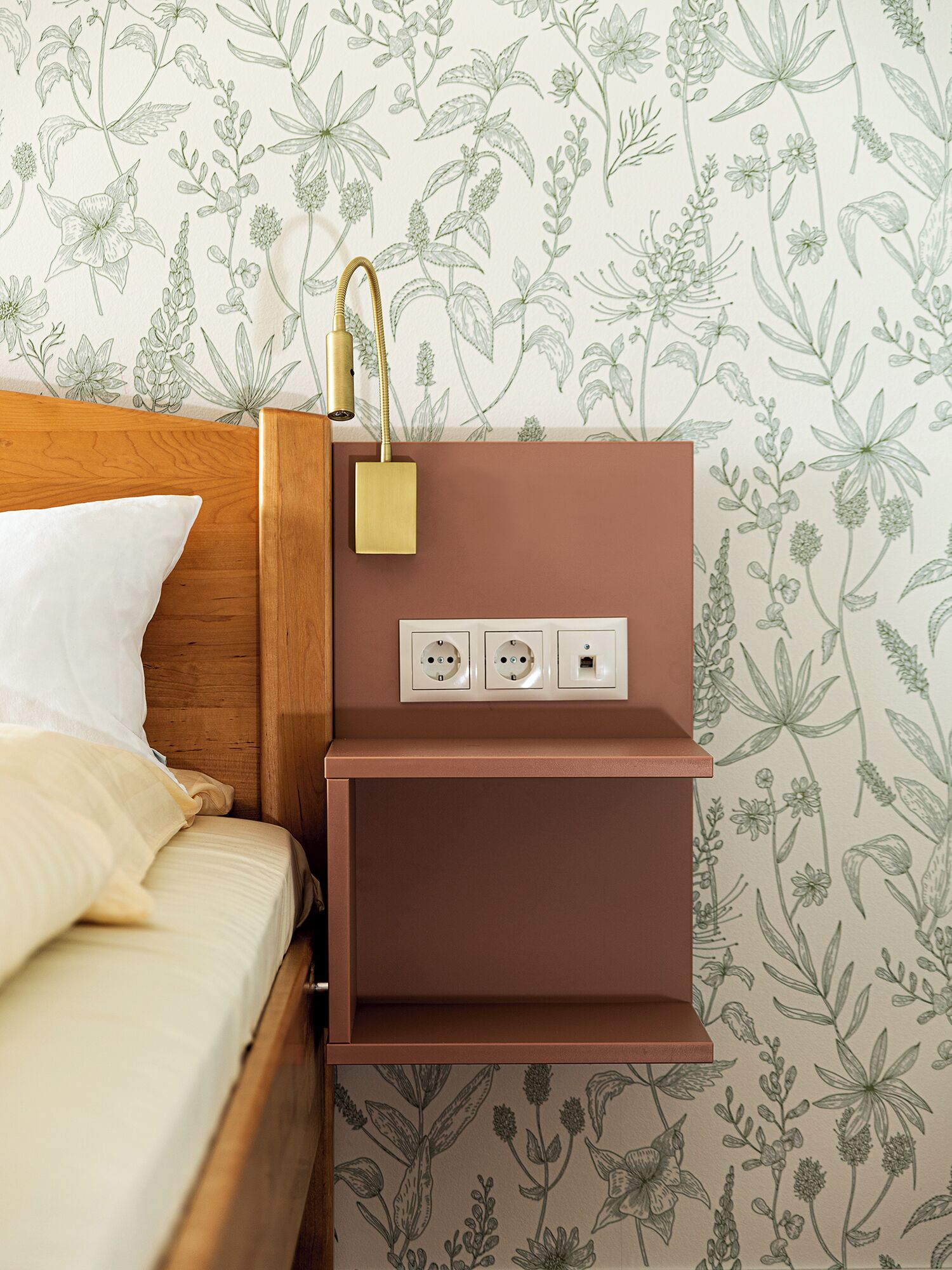 A bed with a wooden headboard, a bedside table with a lamp, and a wall with floral wallpaper.