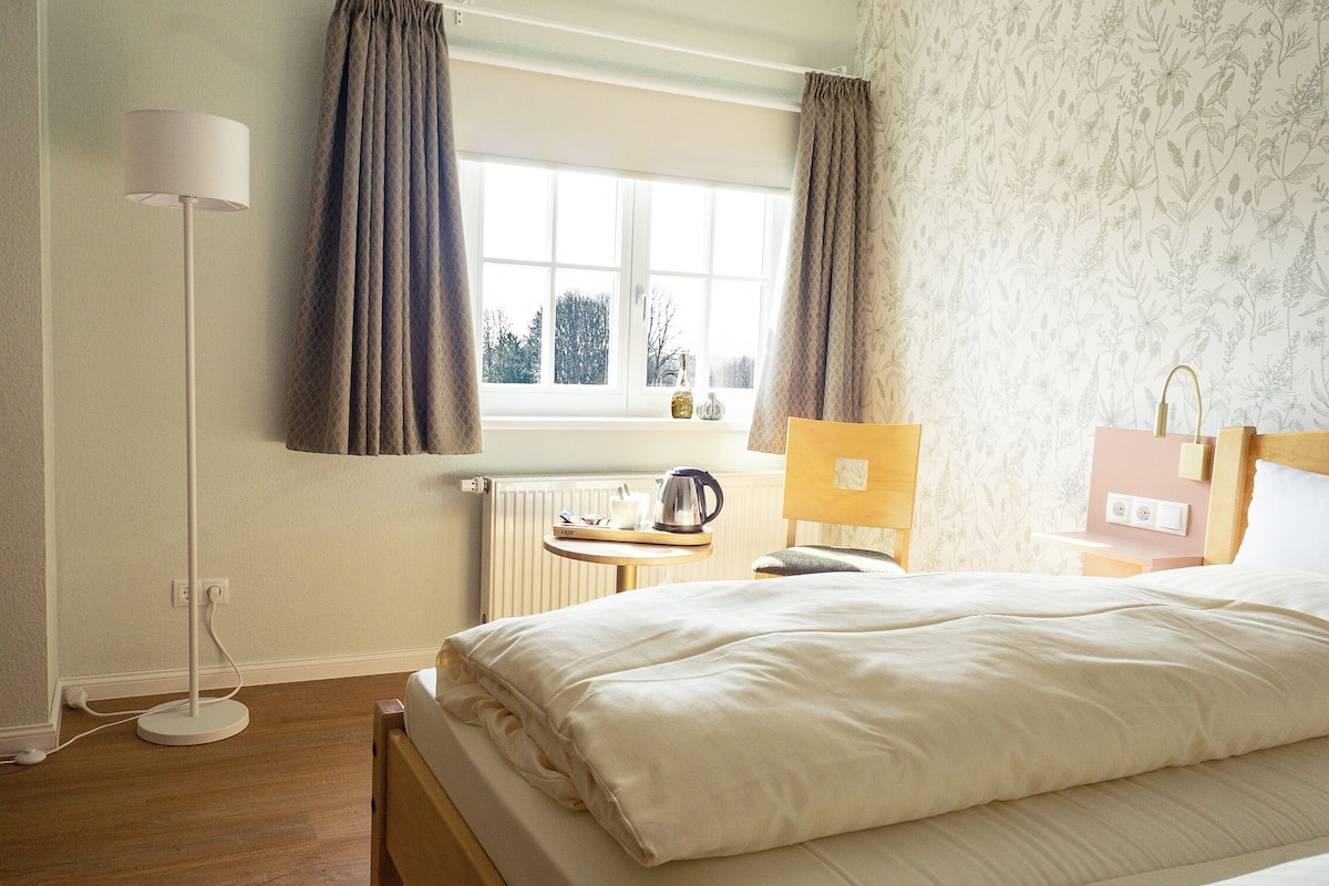 Ein Schlafzimmer mit Bett, Nachttisch mit Lampe, Fenster mit Vorhängen und Heizkörper.