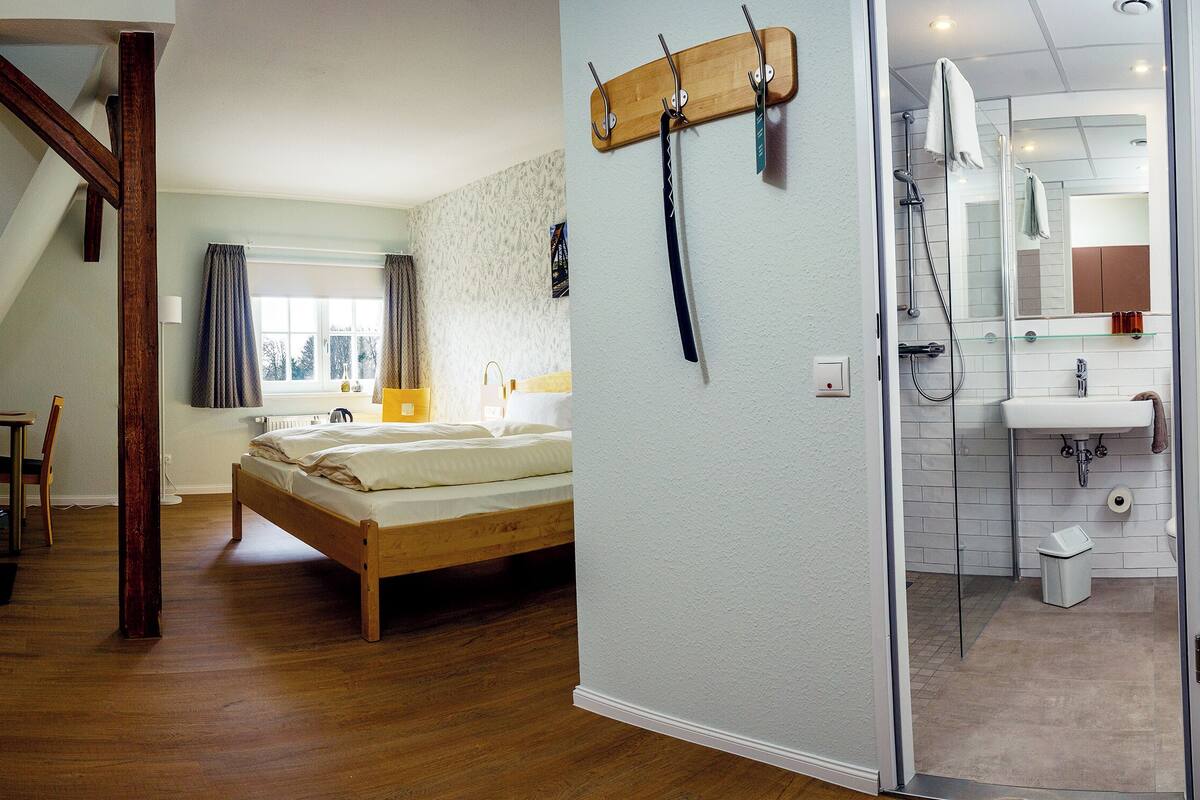 Ein Schlafzimmer mit einem Holzbett, einem Fenster mit Vorhängen, einem Nachttisch und einem an der Wand befestigten Kleiderhaken.