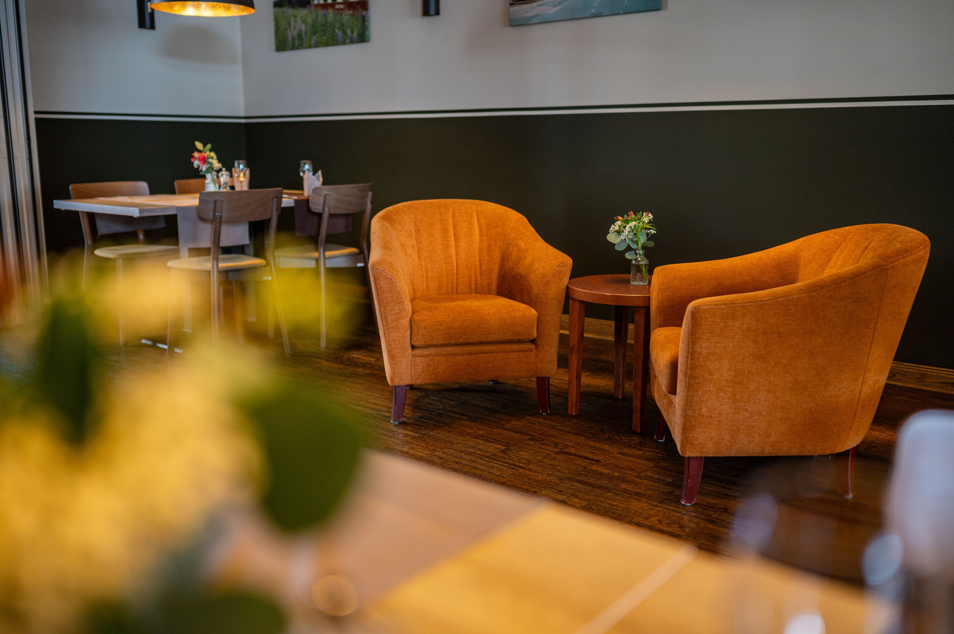 A restaurant interior with wooden flooring, orange armchairs, a small round table with a vase of flowers, and a dining table with chairs.