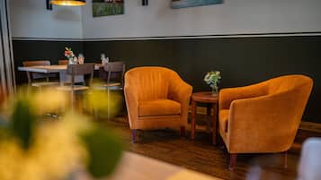 A restaurant interior with wooden flooring, orange armchairs, a small round table with a vase of flowers, and a dining table with chairs.