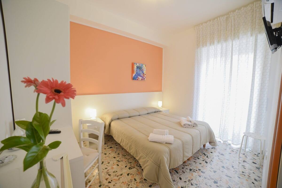 Ein Hotelzimmer mit Bett, Nachttisch, Sessel und einem Blumenstrauß in einer Vase. An der Wand hängt ein Gemälde.