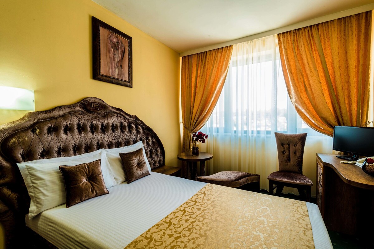 Ein Hotelzimmer mit einem großen Bett, einem Schreibtisch mit Computer, einem Stuhl und einem Fenster mit Vorhängen.