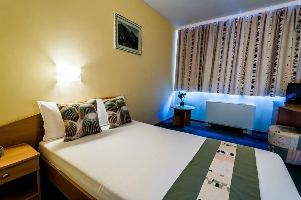 Ein Hotelzimmer mit Bett, Nachttisch, Fenster mit Vorhängen und Heizkörper.