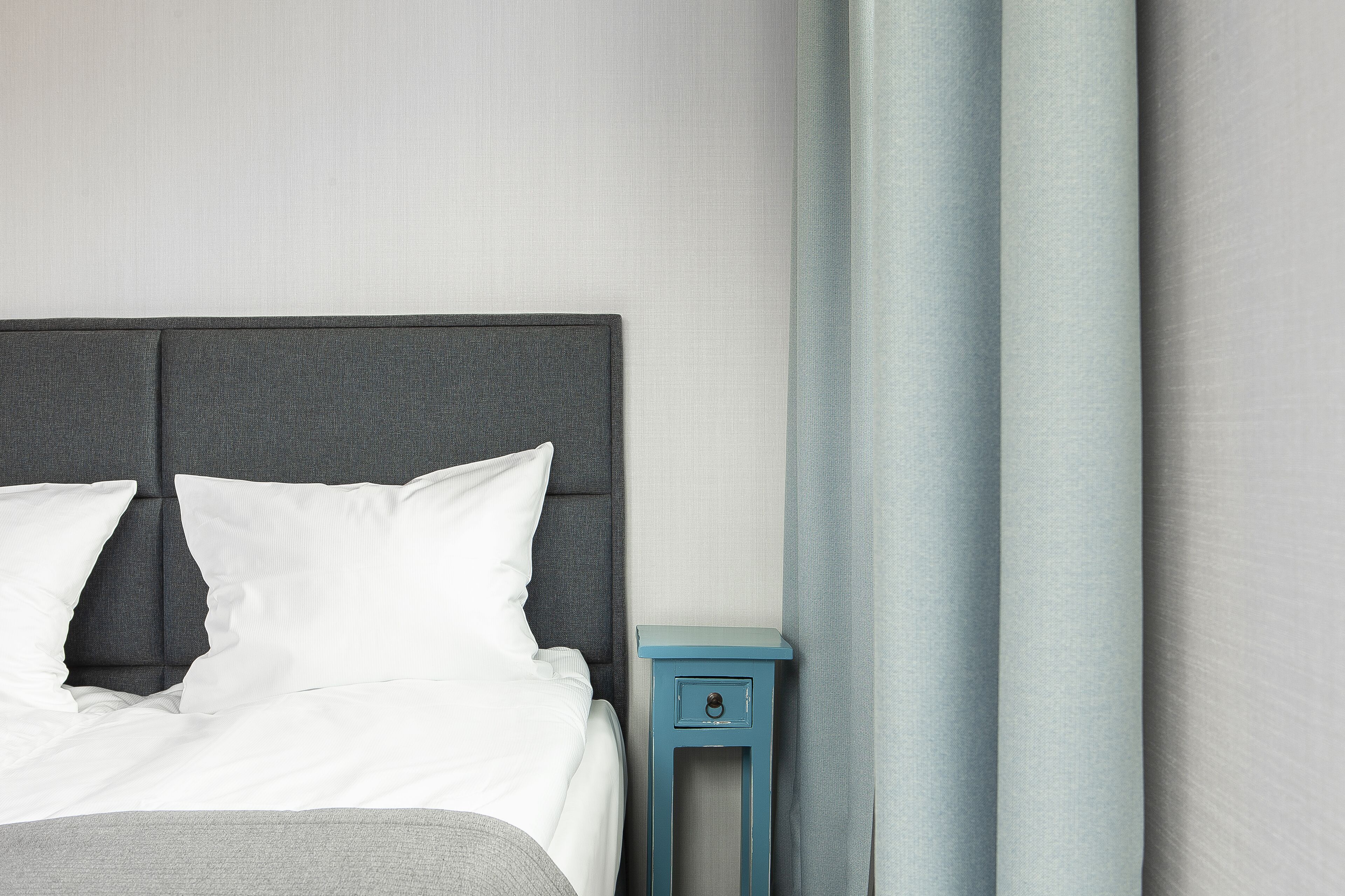 Un lit bien fait, avec une literie et des oreillers blancs, une tête de lit rembourrée de couleur grise et une petite table de chevet bleue dotée d’un tiroir.
