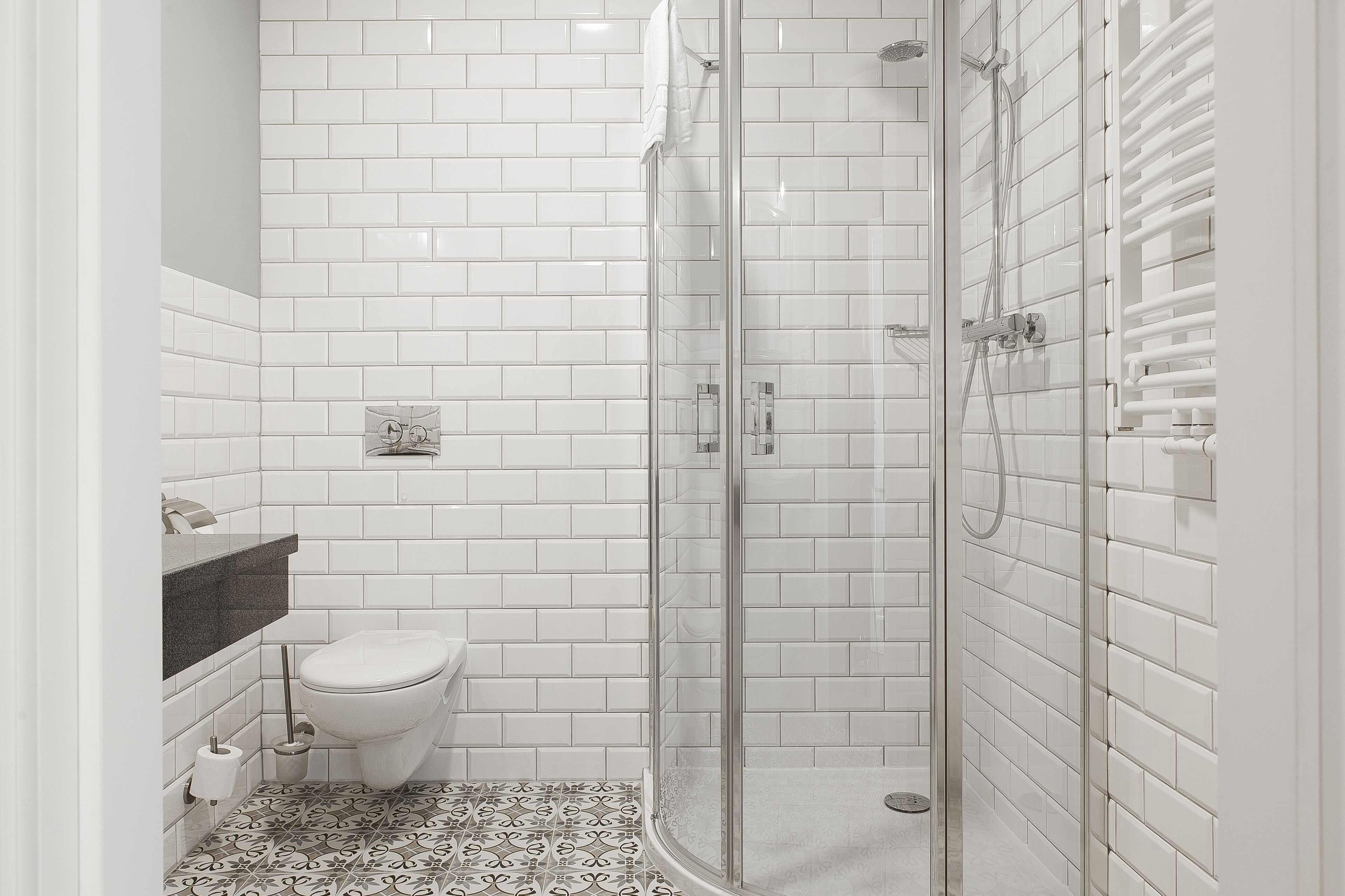 Un baño con azulejos blancos tipo metro, una ducha con mampara de vidrio, un inodoro y un suelo estampado.