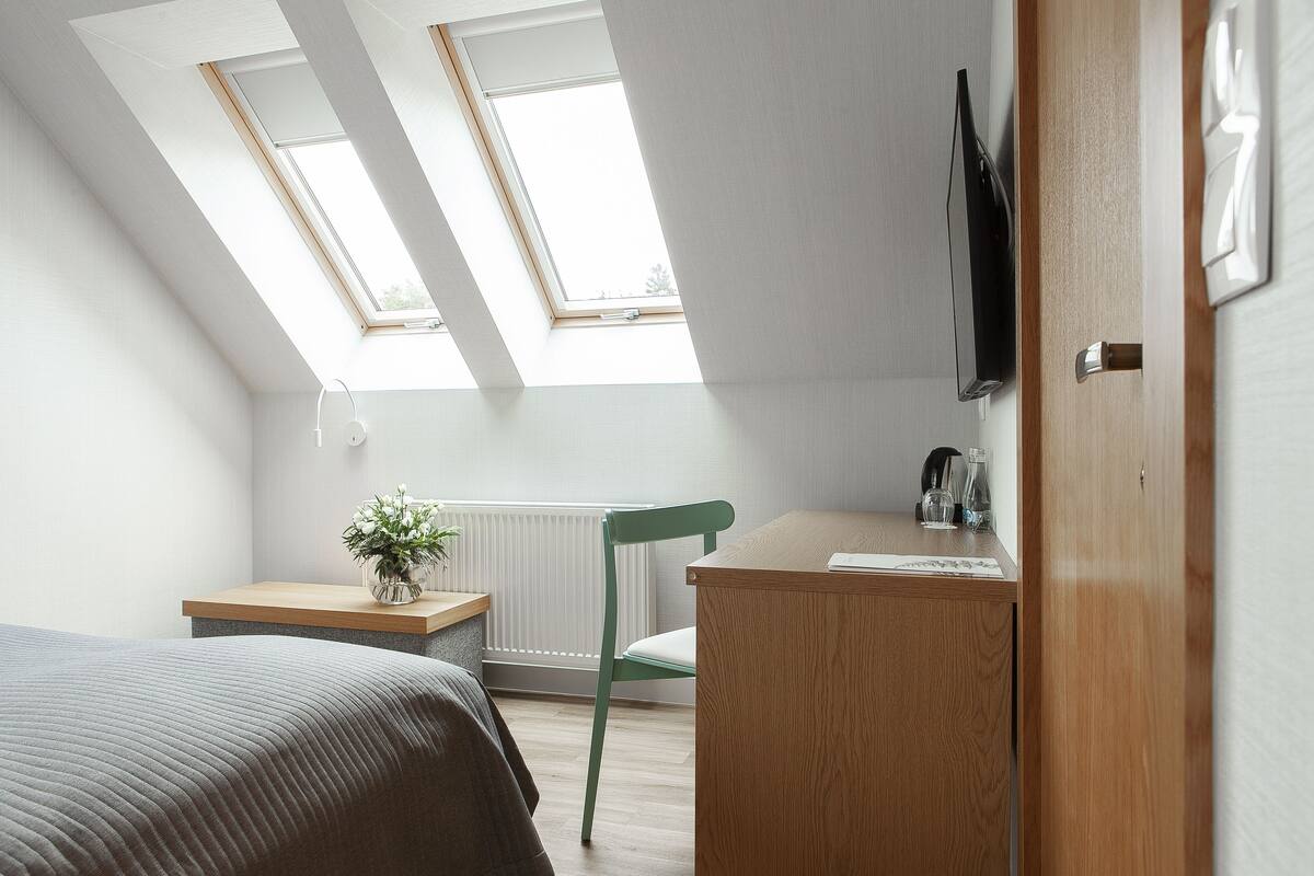 A small, well-lit room with a sloped ceiling, two skylights, a wooden desk, a green chair, and a bed with a gray blanket.