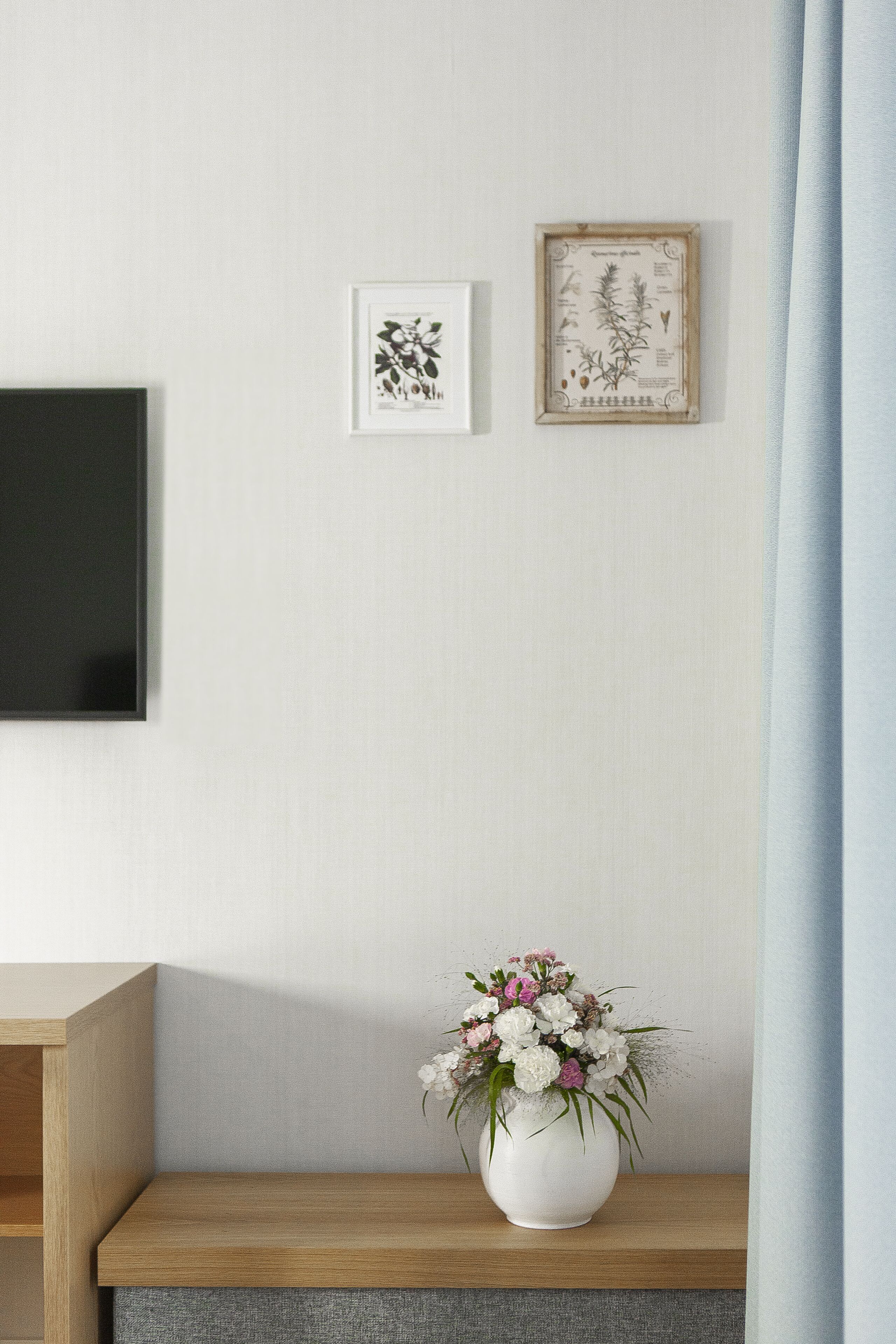A minimalist room with a wooden desk, a white vase with flowers, and framed botanical illustrations on the wall.