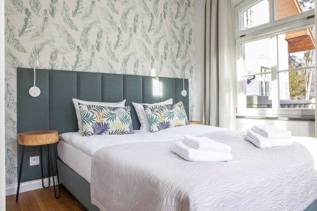 A neatly made bed with a patterned headboard, a bedside table, and a window with daylight.