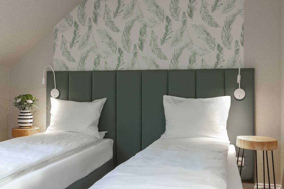 A room with two beds, a green headboard, a wall with a leaf pattern, a bedside table with a plant, and a small wooden stool.
