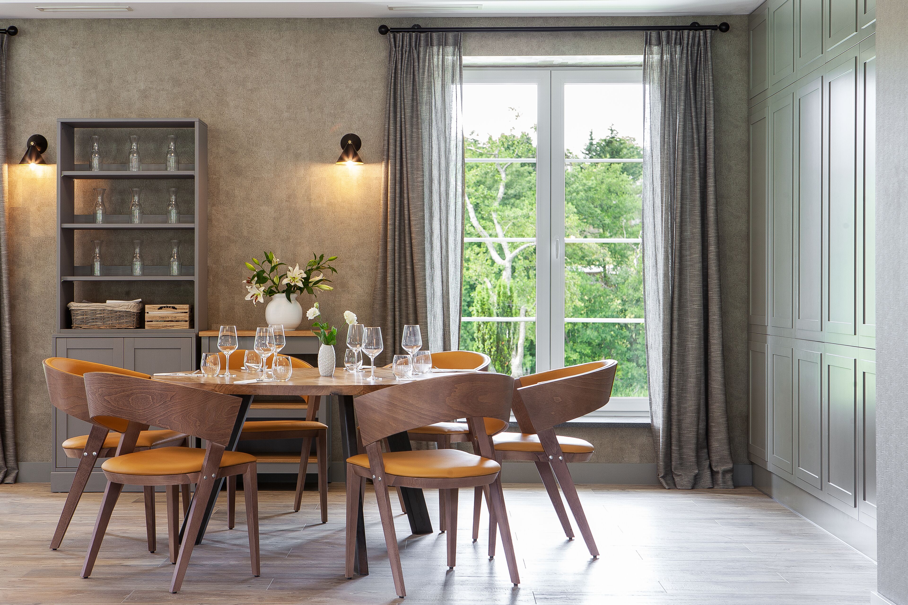 A dining area with a round table, six wooden chairs, a bookshelf, and a window with curtains.