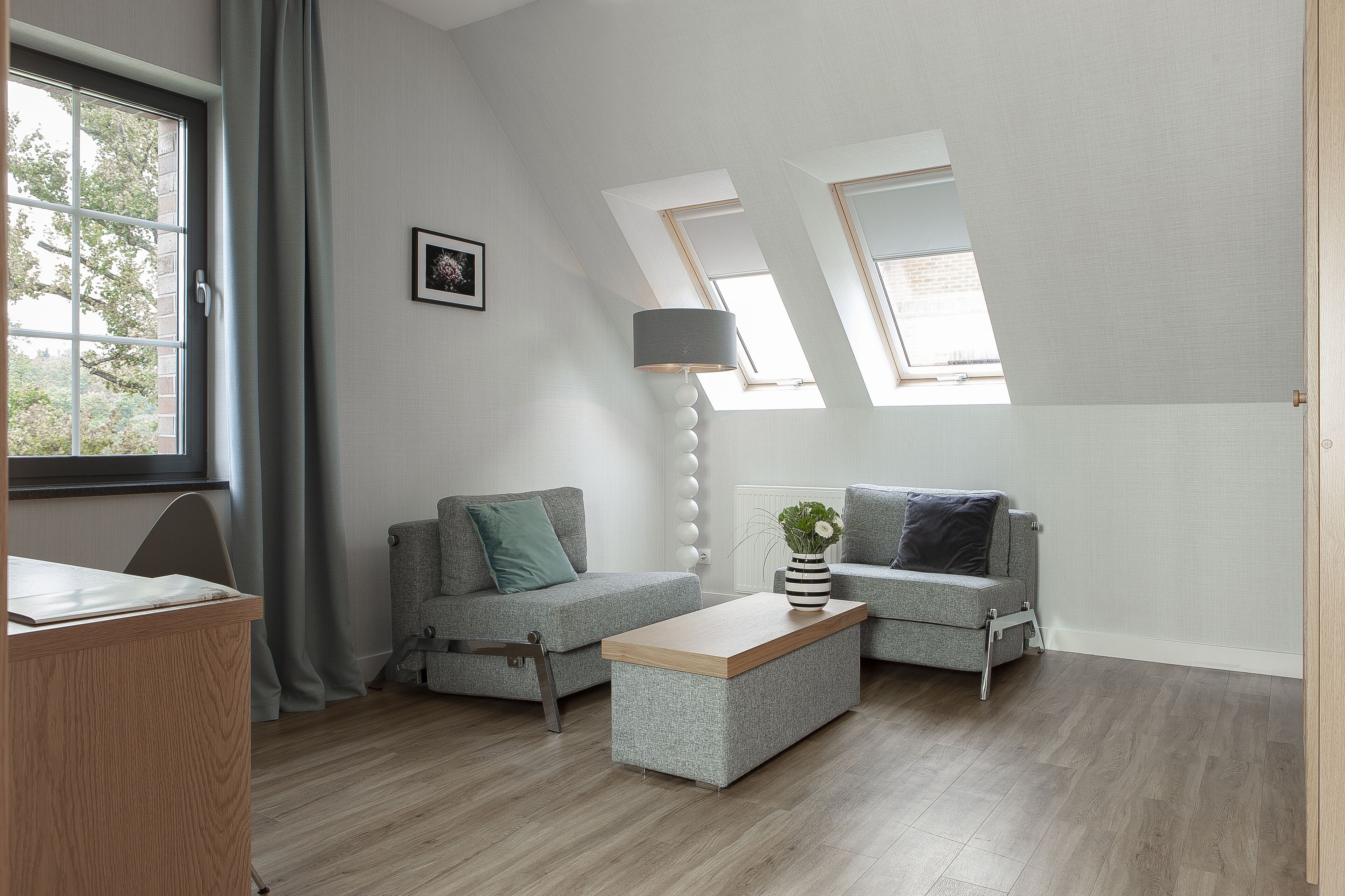 A modern living room with two grey sofas, a wooden coffee table, and a lamp. There are skylights on the ceiling and a window with curtains.