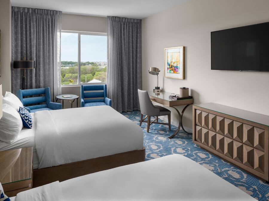 A hotel room with two beds, a desk, a chair, a TV, and a view of the outdoors.