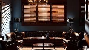 Sala de estar com decoração escura, incluindo um sofá de couro, uma mesa de centro redonda e uma grande janela com persianas.